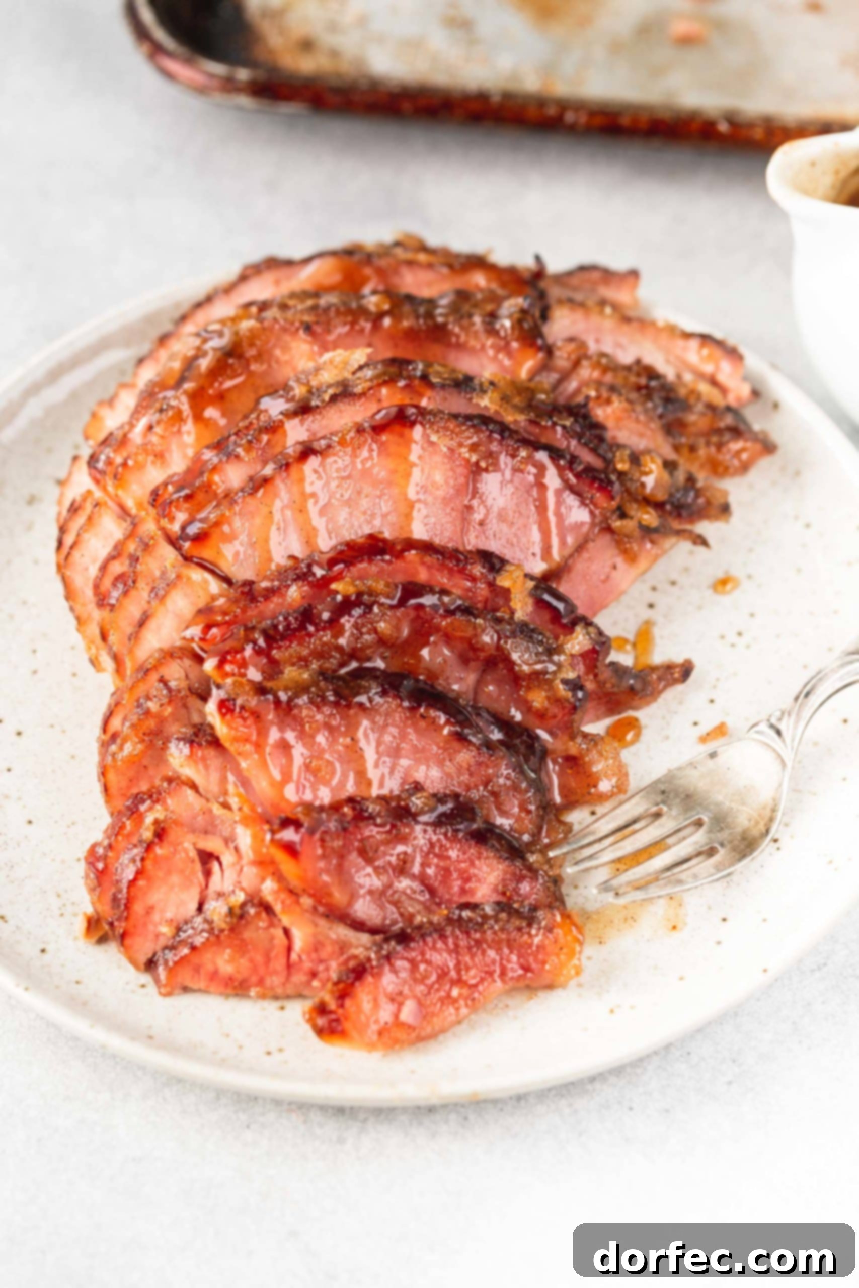 A spiral cut honey glazed ham on a serving plate with a fork next to it. The slices are slightly separated so you can see how juicy the ham is and how beautiful the glaze is.