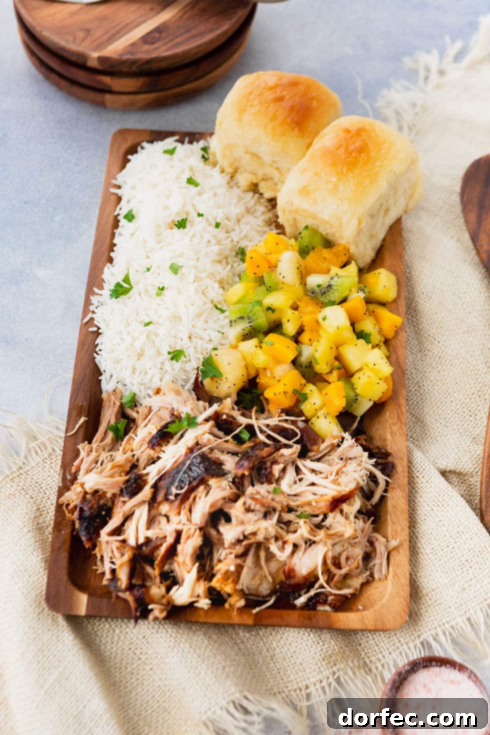 A photo of a rectangular wooden platter full of kalua pulled pork, herbed rice, tropical fruit salad, and golden Hawaiian rolls.