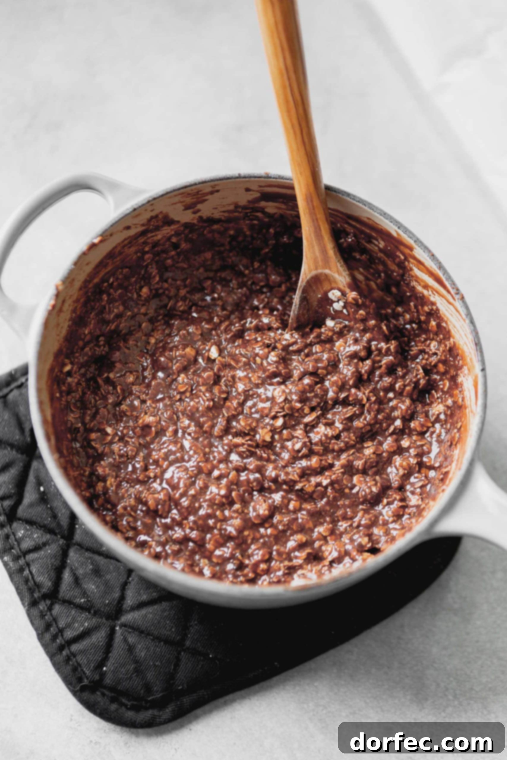 A saucepan containing the fully mixed no-bake cookie dough, a blend of oatmeal and chocolate, with a wooden spoon resting inside.