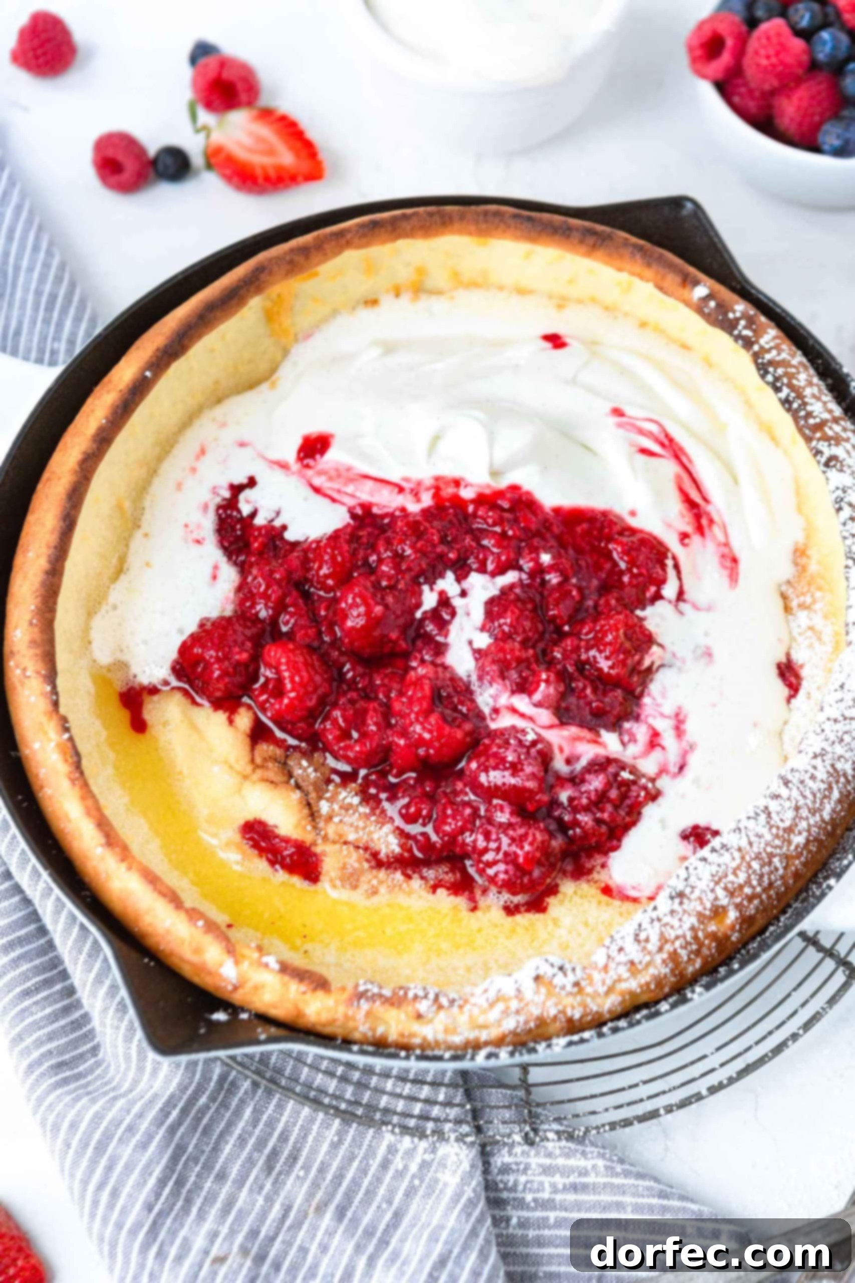 Dutch Baby Pancake A freshly baked Dutch Baby Pancake, golden and puffed, topped with swirls of whipped cream and a colorful assortment of fresh strawberries, blueberries, and raspberries. It sits in a rustic cast iron skillet, ready to be served.