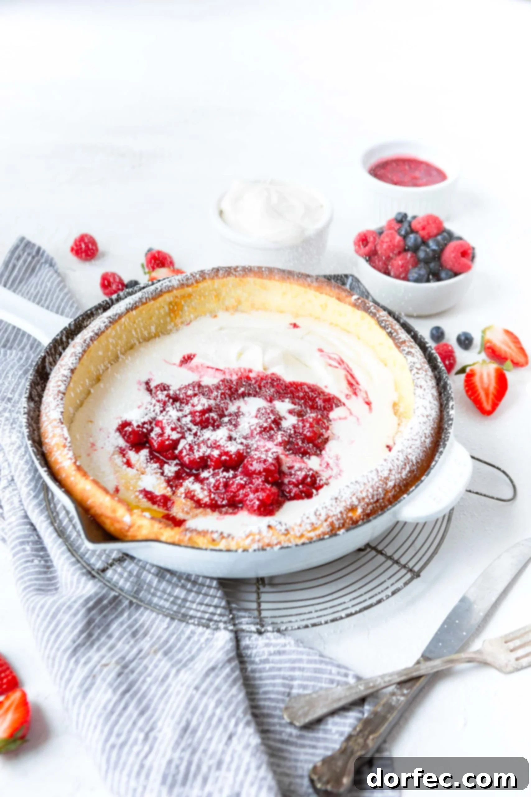 Dutch Baby Pancake A close-up view of a baked Dutch Baby Pancake in a cast iron skillet, beautifully puffed and golden. It is elegantly garnished with fresh whipped cream, a scattering of mixed berries (strawberries, blueberries, raspberries), and a dusting of powdered sugar, highlighting its appealing textures.