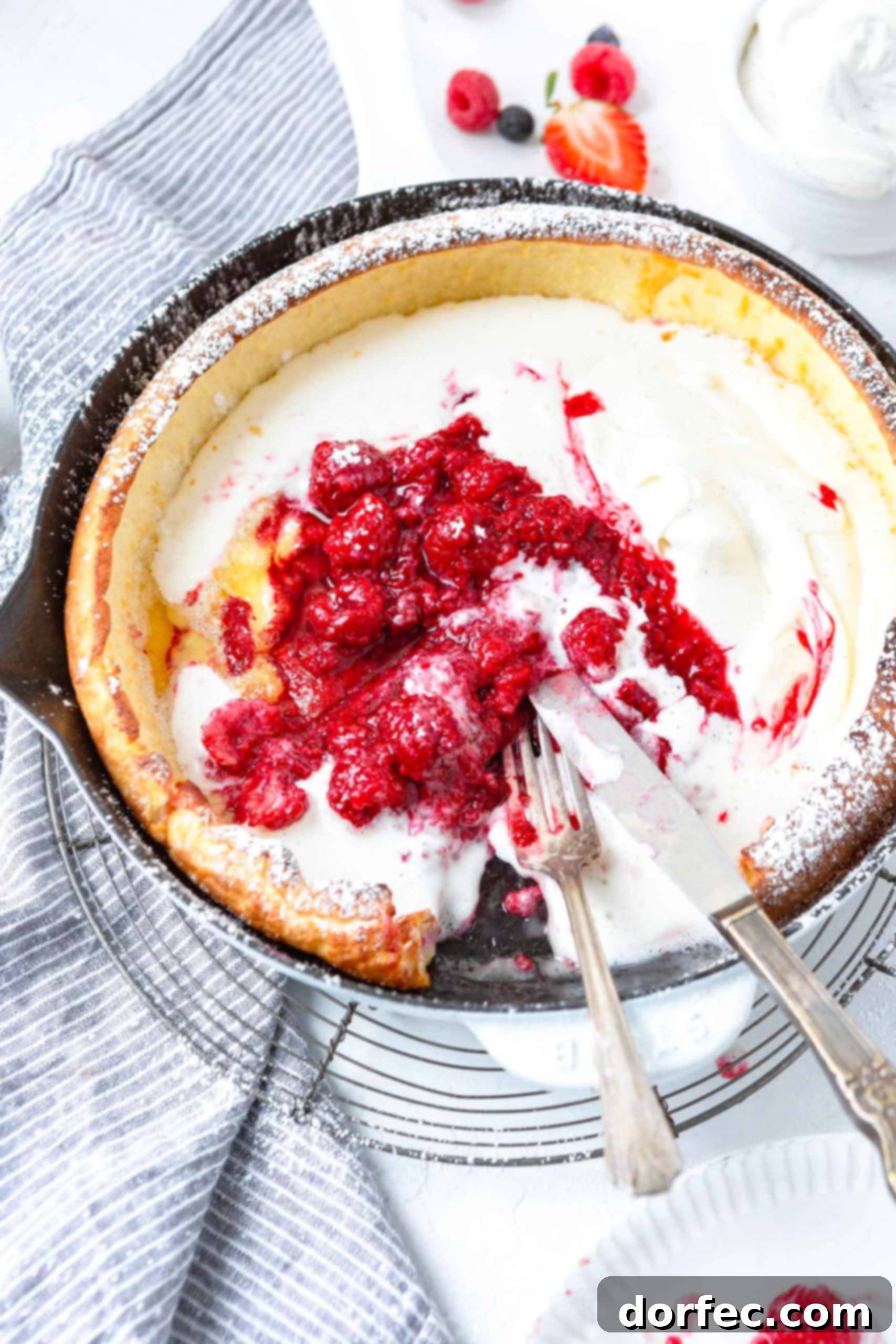 Dutch Baby Pancake A Dutch Baby Pancake, golden and beautifully puffed, resting in a cast iron skillet. A slice has been removed, revealing its custardy interior. A fork and knife lie elegantly beside the pan, ready for serving.