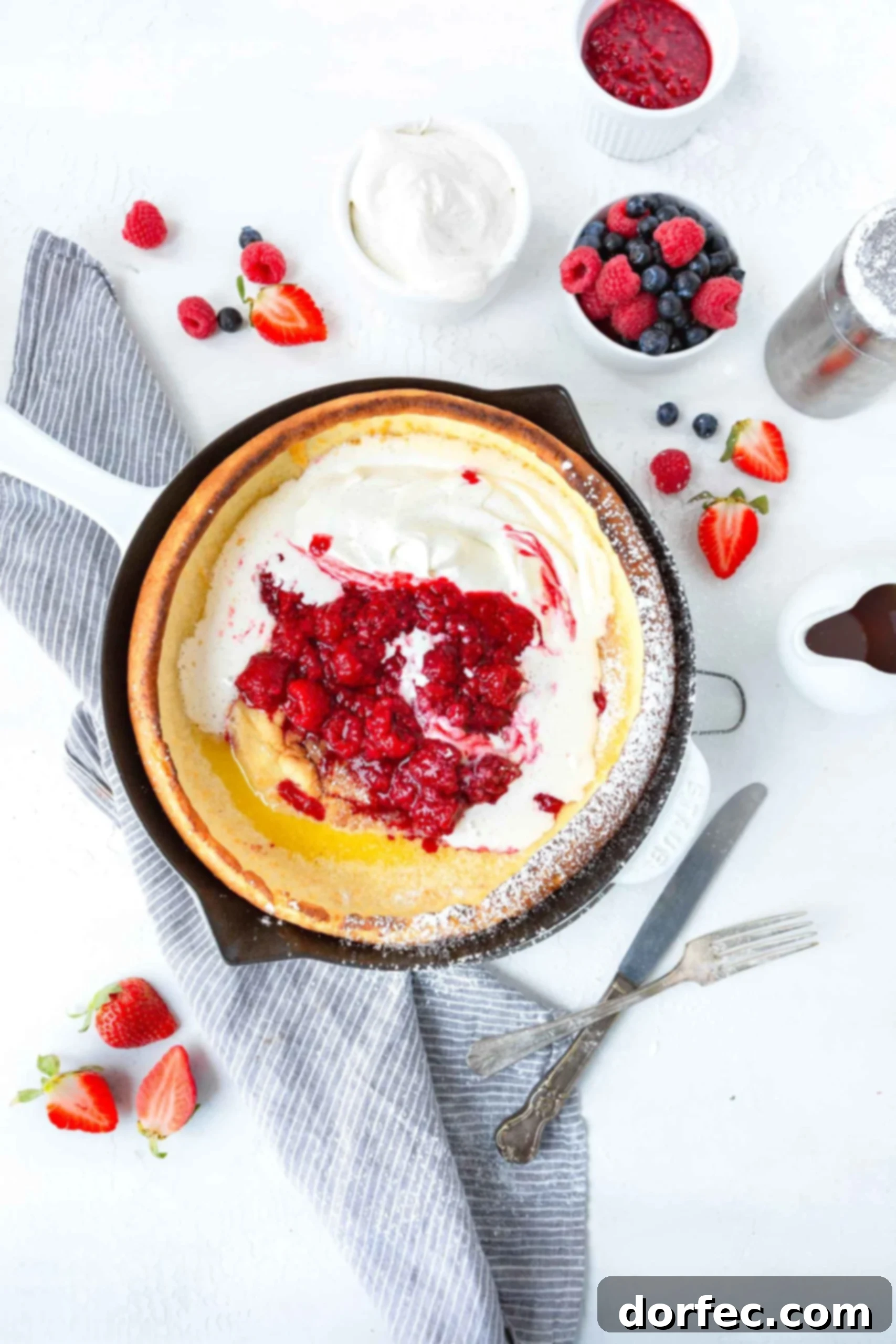 Dutch Baby Pancake A beautifully baked Dutch Baby Pancake in a cast iron skillet, generously topped with whipped cream and an abundance of fresh, colorful berries. Beside it, a small cup overflows with extra fresh berries, ready to be added, creating a vibrant breakfast scene.