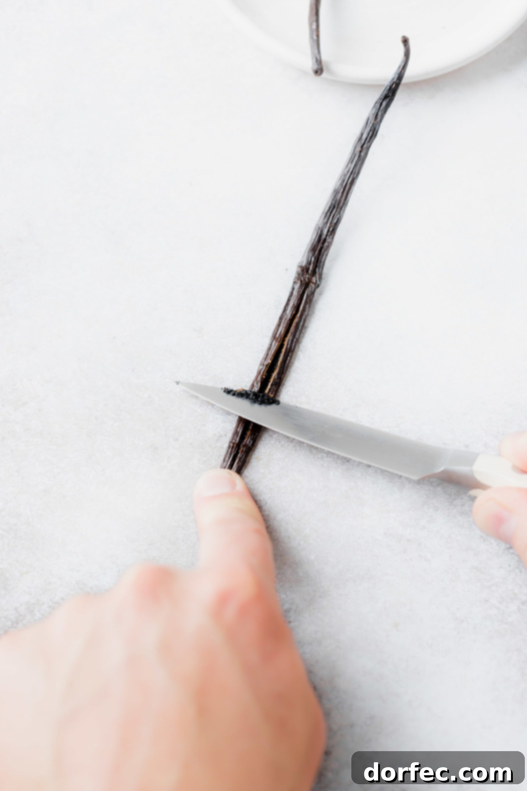 A skilled hand demonstrates how to scrape out the aromatic seeds from a vanilla bean pod using a sharp paring knife on a wooden cutting board.