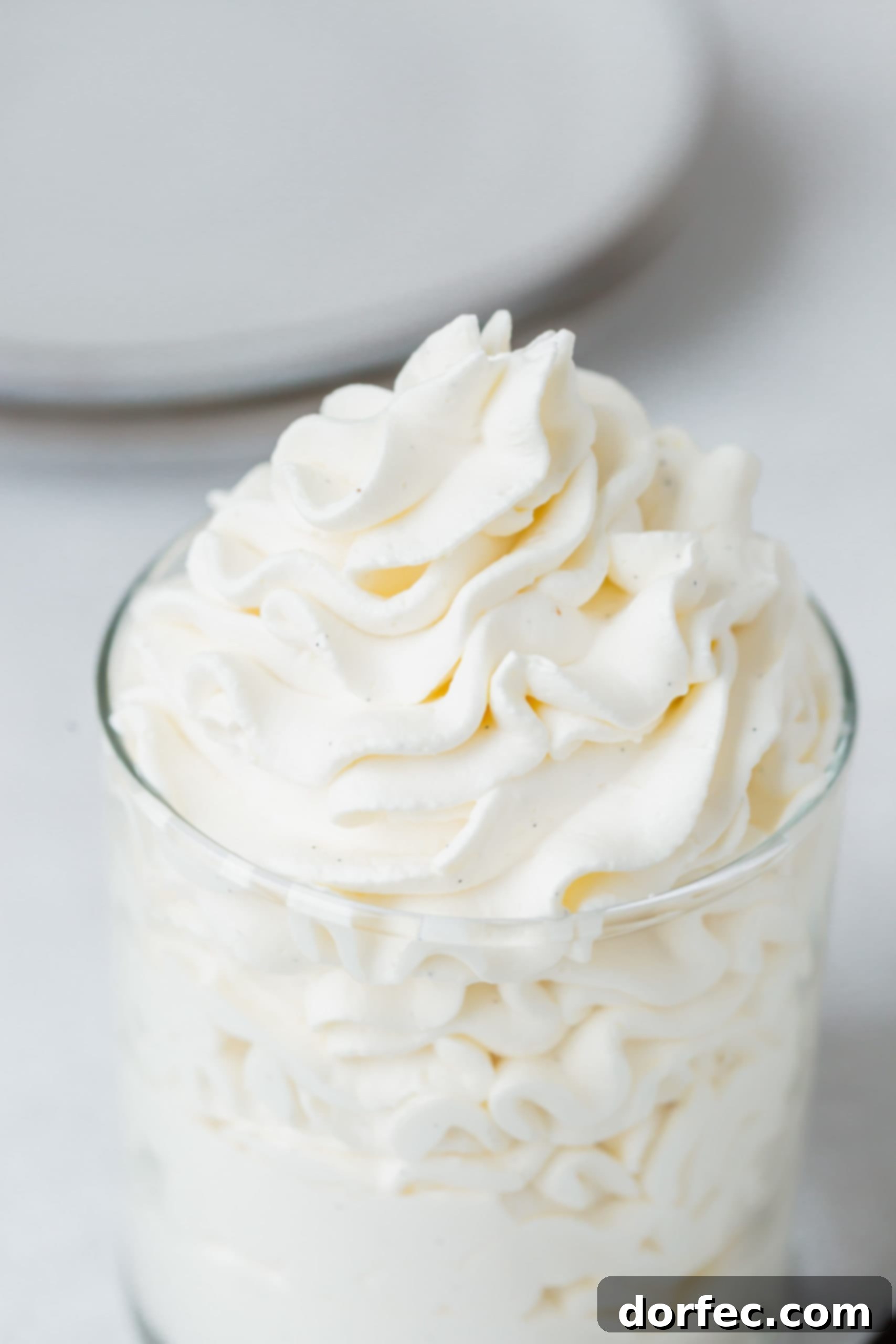 A close-up view of a glass filled with exquisitely smooth and airy vanilla bean whipped cream, topped with a single, dark vanilla bean specks visible.