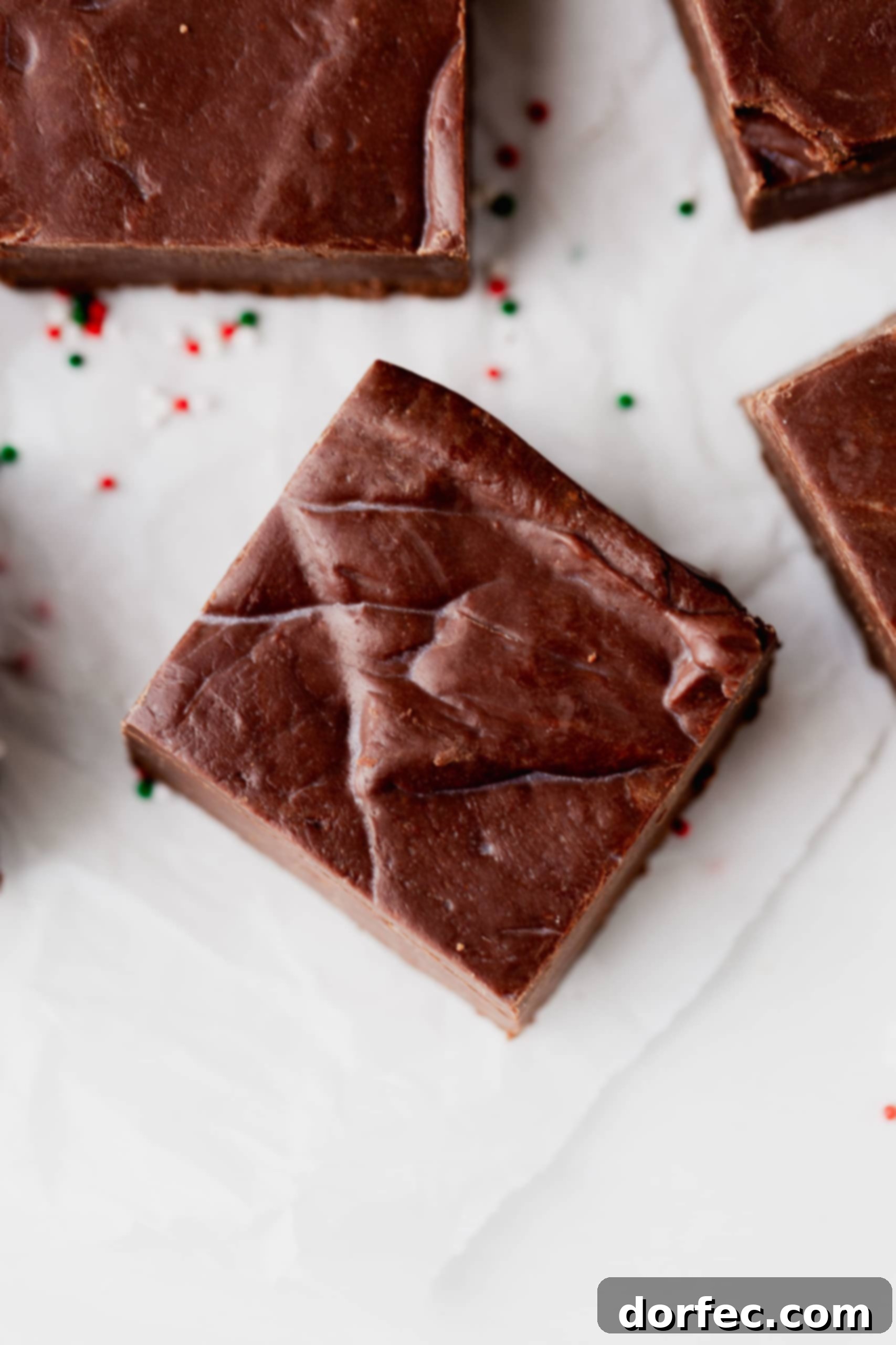 A close-up, top-down view of a single square piece of rich chocolate fudge, perfectly cut and ready to enjoy. Other pieces of fudge are visible in the background, creating an enticing display.