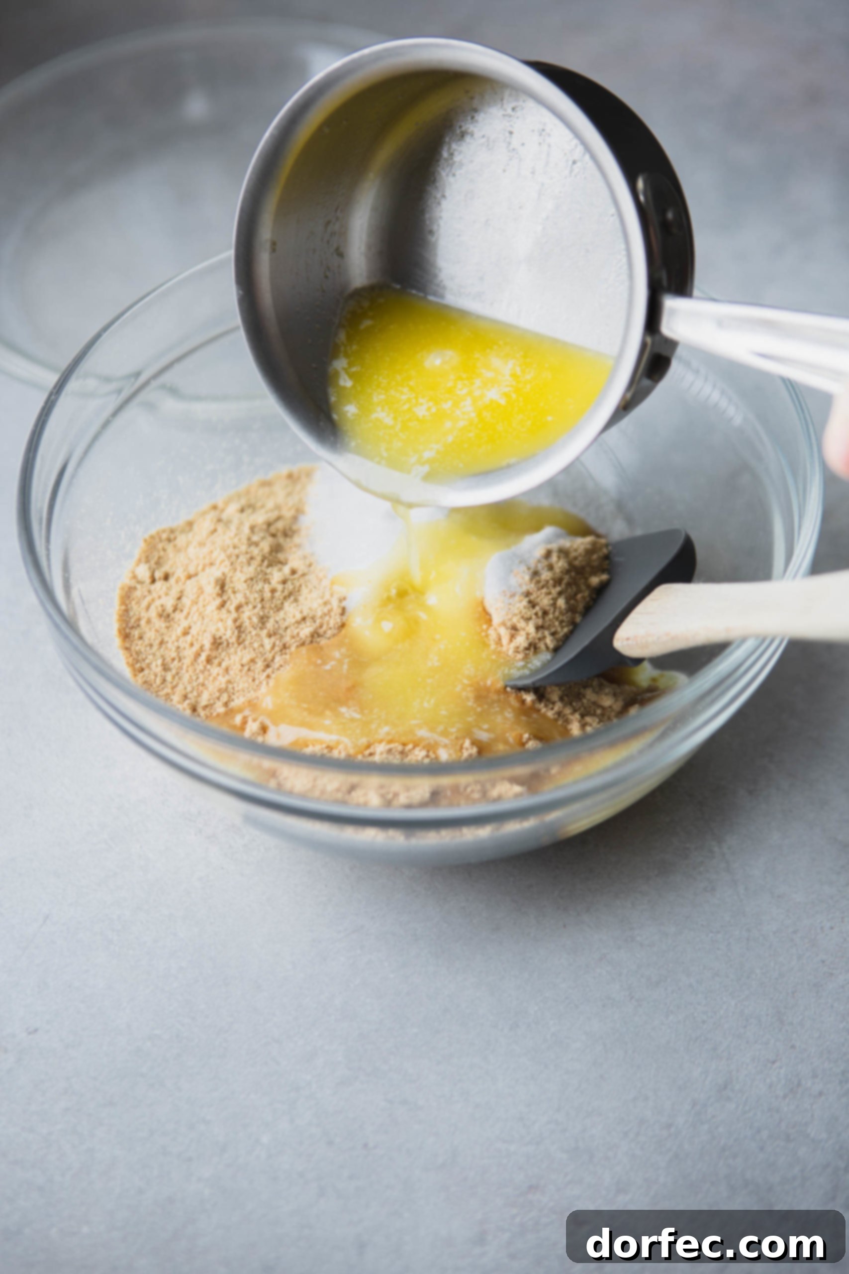 Graham Cracker Crust Melted butter is being poured over a mixture of graham cracker crumbs and sugar in a clear glass mixing bowl, initiating the process of creating a perfect pie crust.