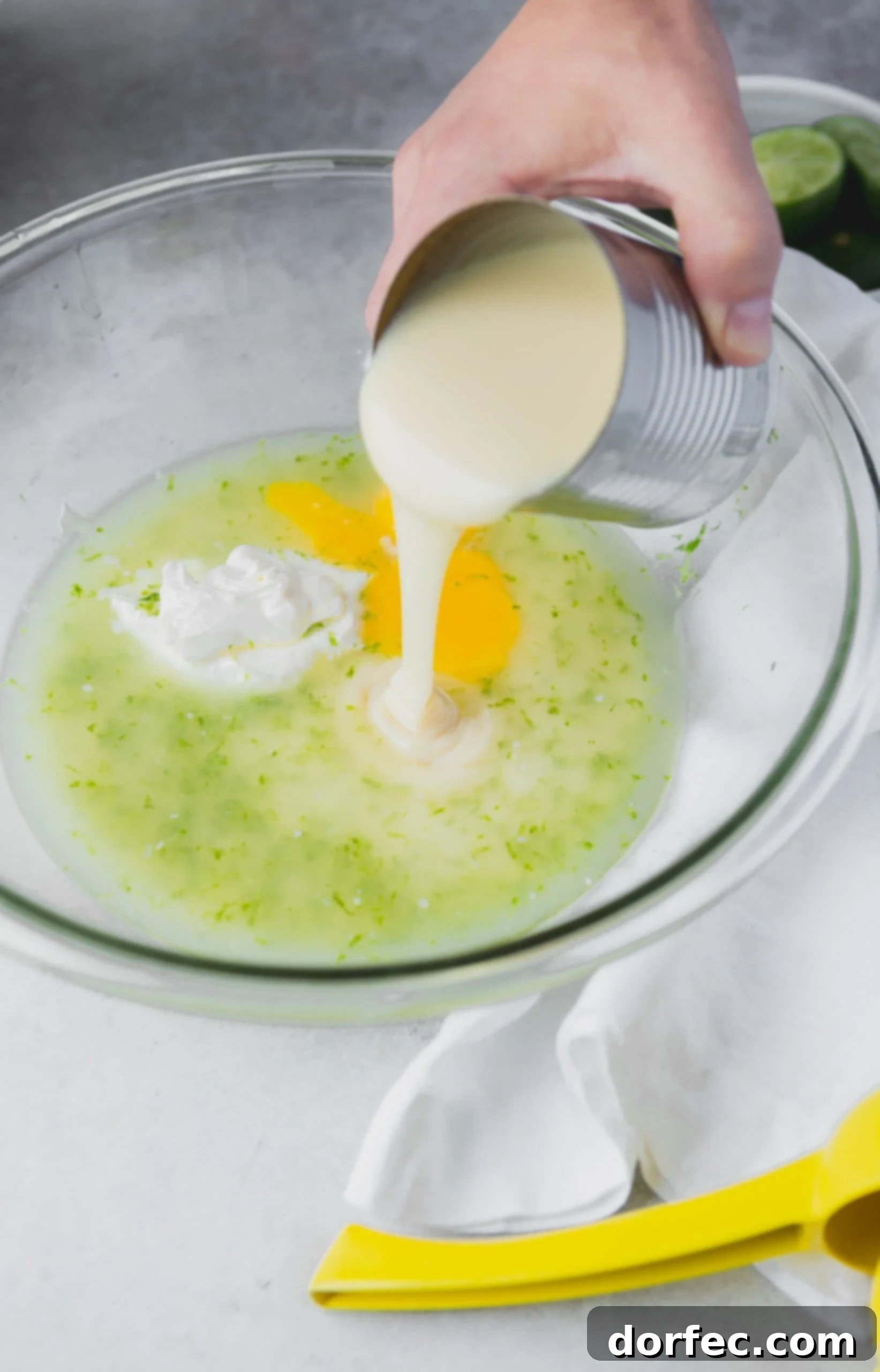 Key Lime Pie Filling Preparation Rich, creamy sweetened condensed milk being carefully poured from a can into a mixing bowl, joining egg yolks, fresh lime juice, vibrant lime zest, and cool sour cream, ready to be whisked into the perfect Key Lime Pie filling.