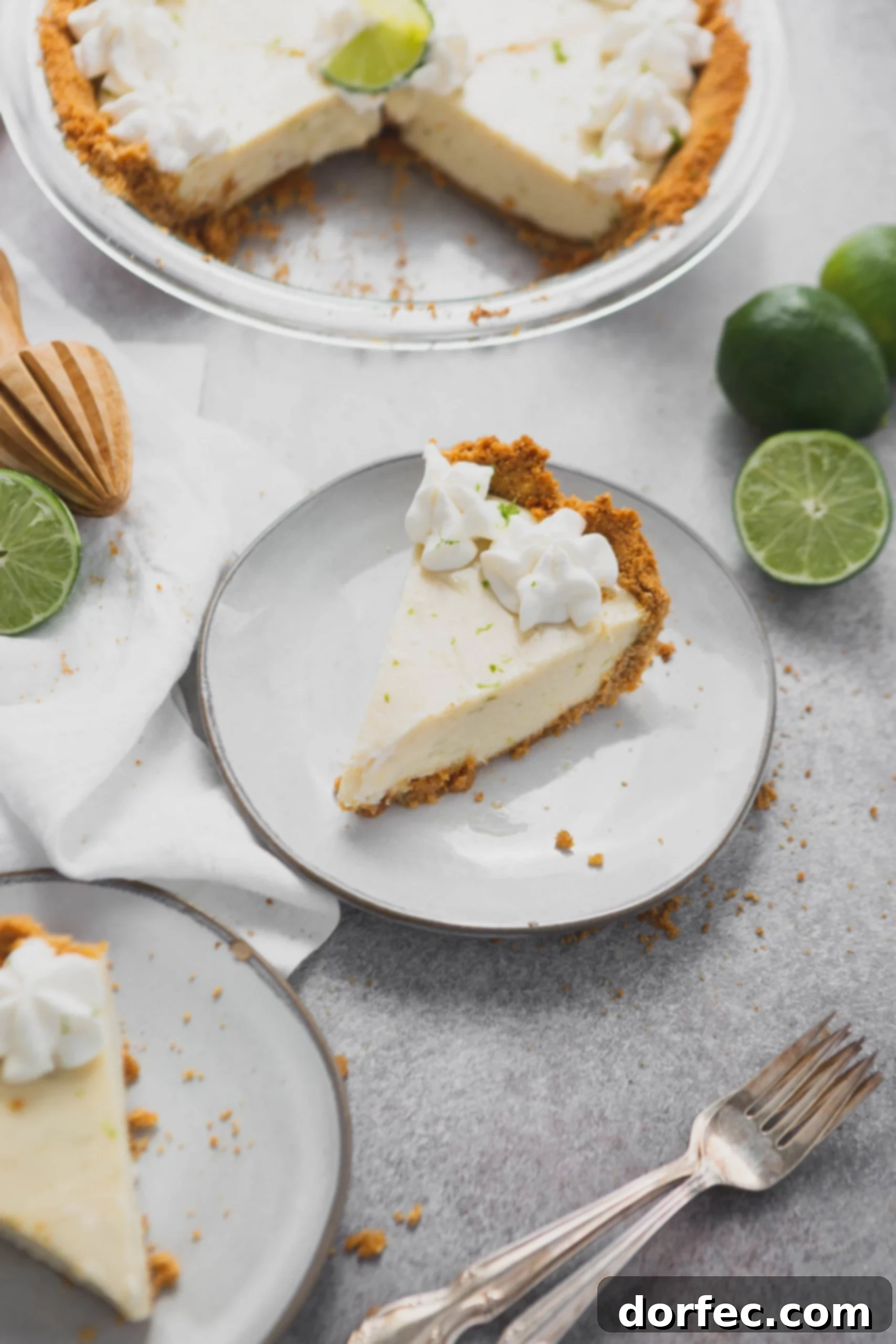 Slice of Key Lime Pie A beautiful slice of Key Lime Pie presented on a white dessert plate, featuring its graham cracker crust, a generous layer of creamy Key lime filling, and two delicate dollops of whipped cream on top. A halved lime and another slice of pie rest beside it, inviting indulgence.