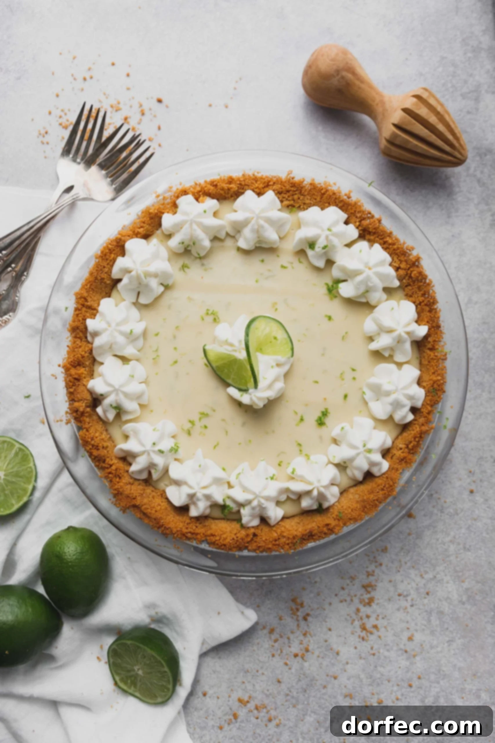 Whole Key Lime Pie A beautifully presented Key Lime Pie, whole and in its graham cracker crust, adorned with elegant dollops of whipped cream around the edge and two fresh lime slices in the center. Fresh limes and forks are artfully arranged beside the pie, ready for serving.
