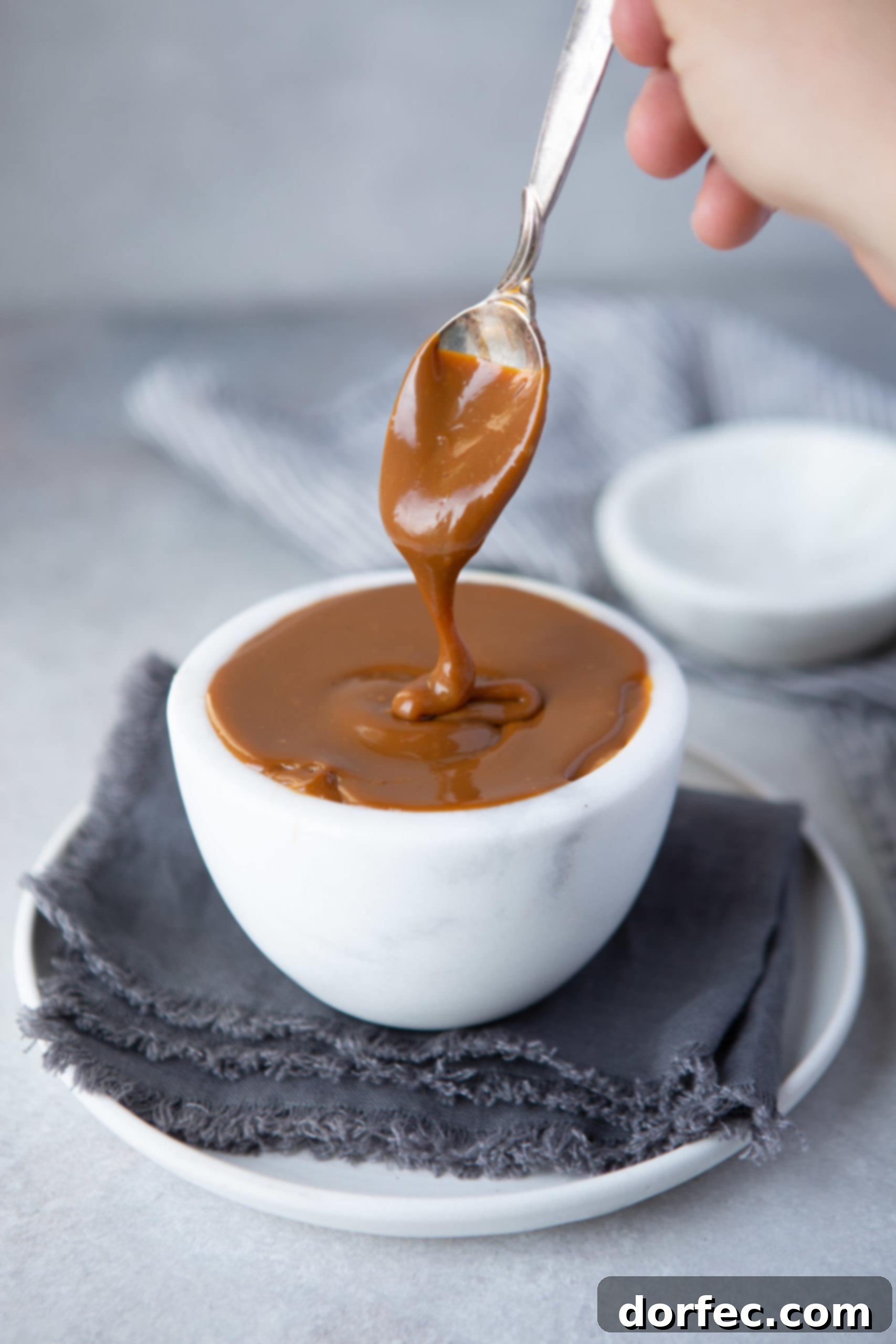 A close-up photo of a white cup brimming with luscious homemade dulce de leche, with a spoon gently lifting some, creating a rich, golden drizzle.