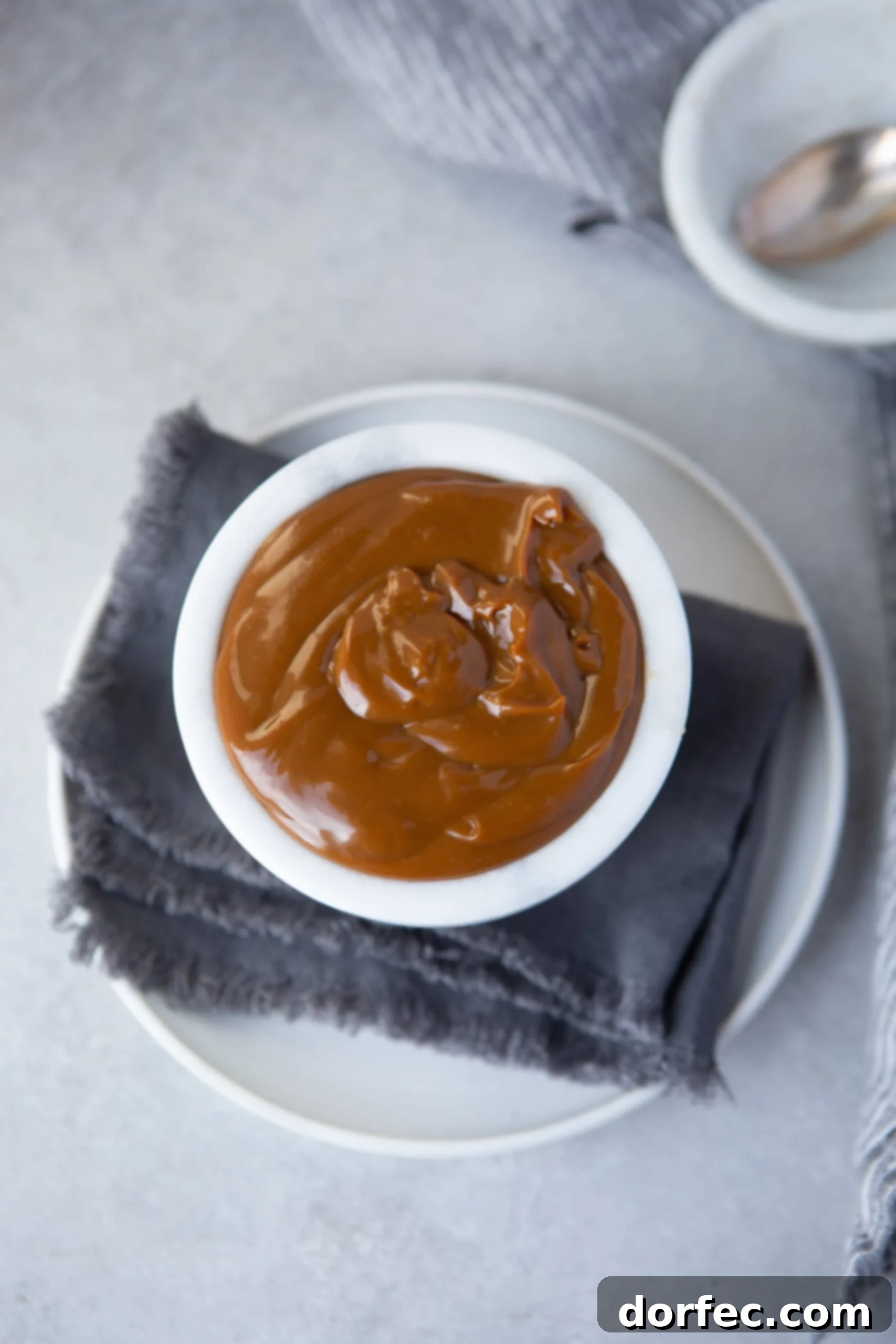 An overhead shot of a white ceramic cup filled with creamy dulce de leche, resting on a blue cloth napkin and a white plate, highlighting its smooth texture.