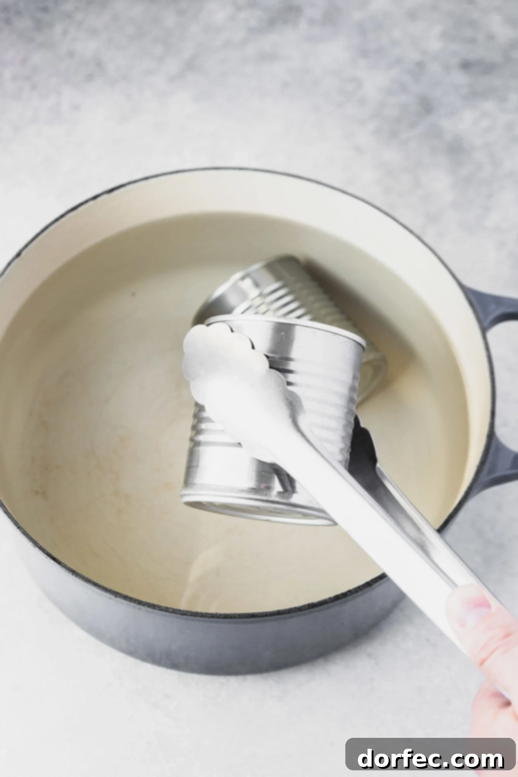 A pair of metal tongs carefully removing a closed can of cooked sweetened condensed milk from a pot of simmering water on the stovetop.