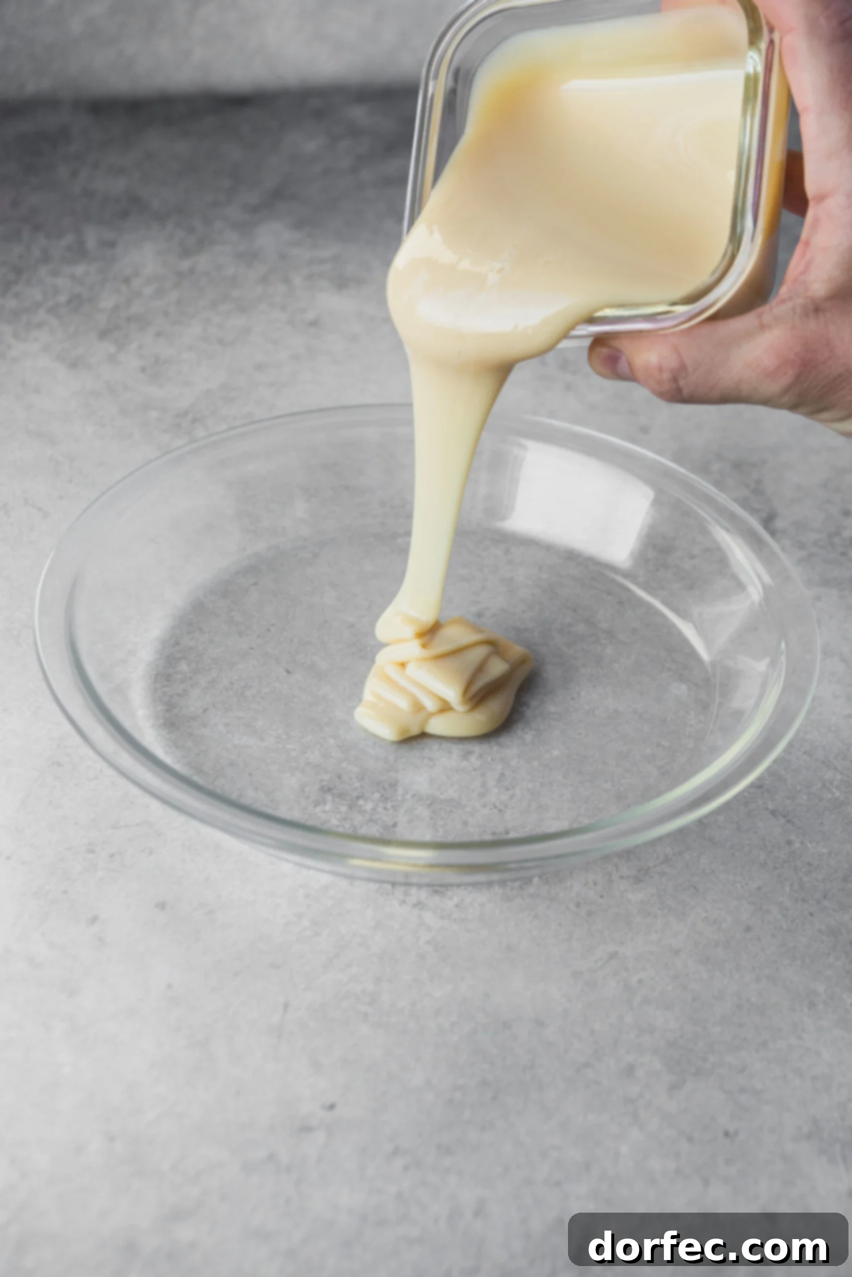 Sweetened condensed milk being poured from a can into a clear glass pie plate, showcasing its thick, creamy texture before transformation.