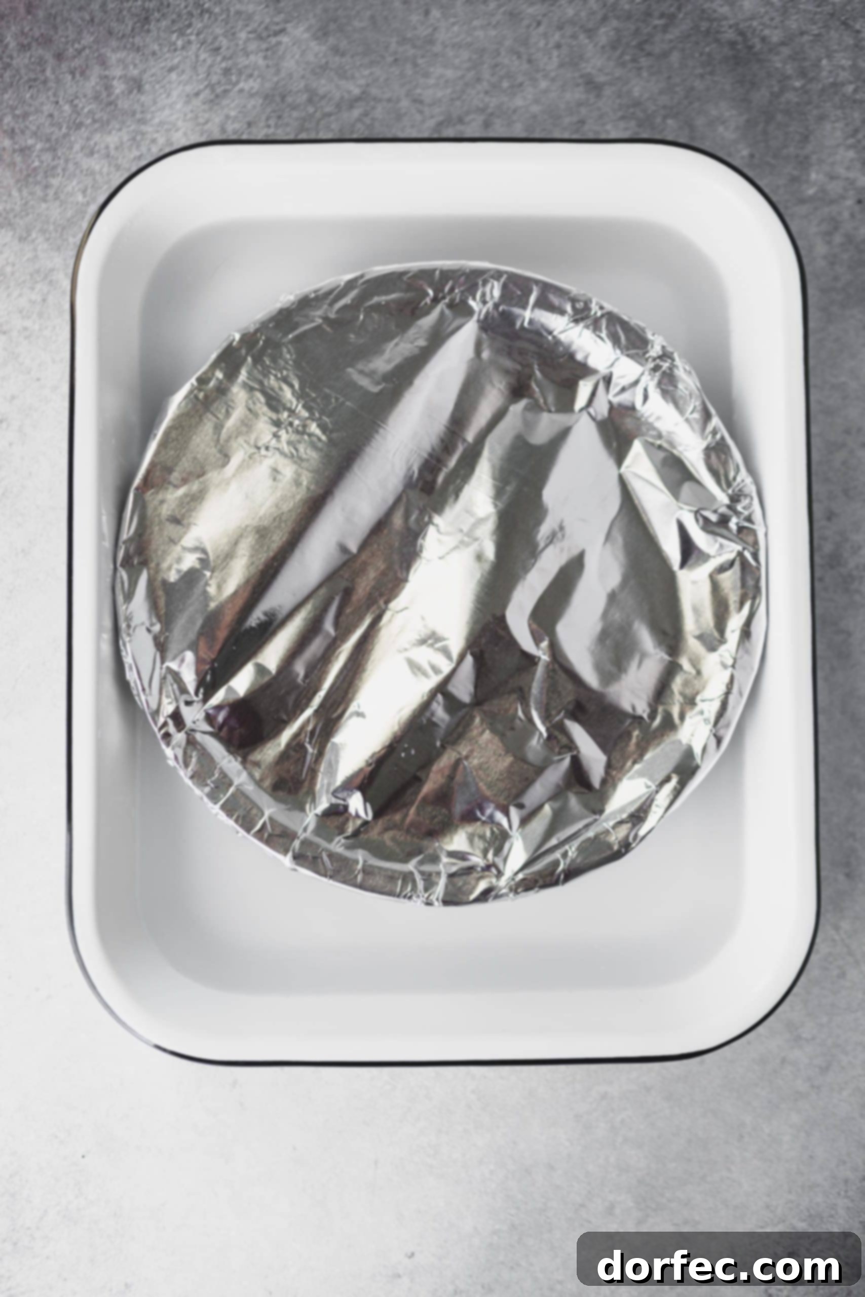 An oven-safe pie plate, tightly covered with aluminum foil, sitting in a larger casserole dish filled halfway with water, prepared for the oven method of making dulce de leche.