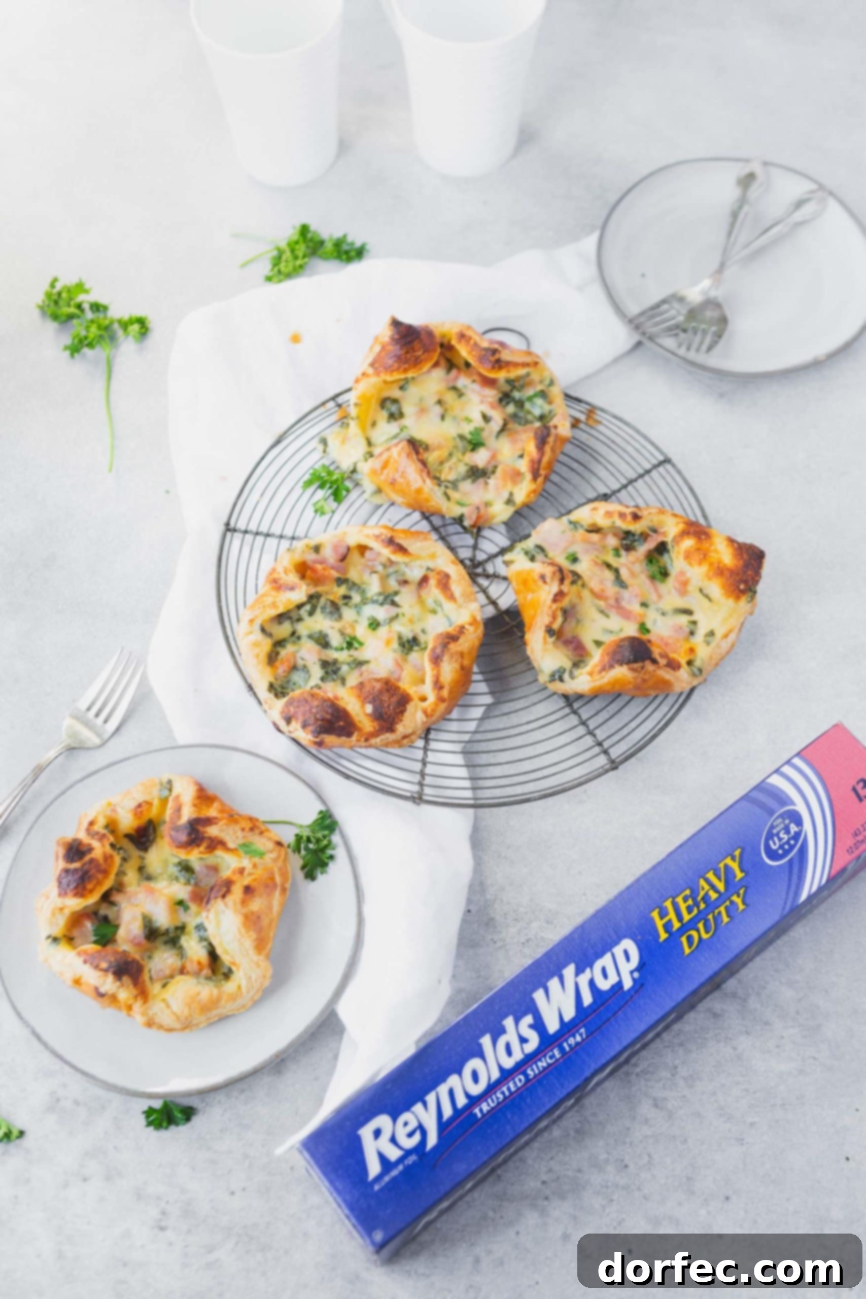 Three golden ham and cheese puffs on a circular cooling rack, with one puff on a white plate, all next to a box of Reynolds Wrap Heavy Duty foil, showcasing a delicious and easy treat.