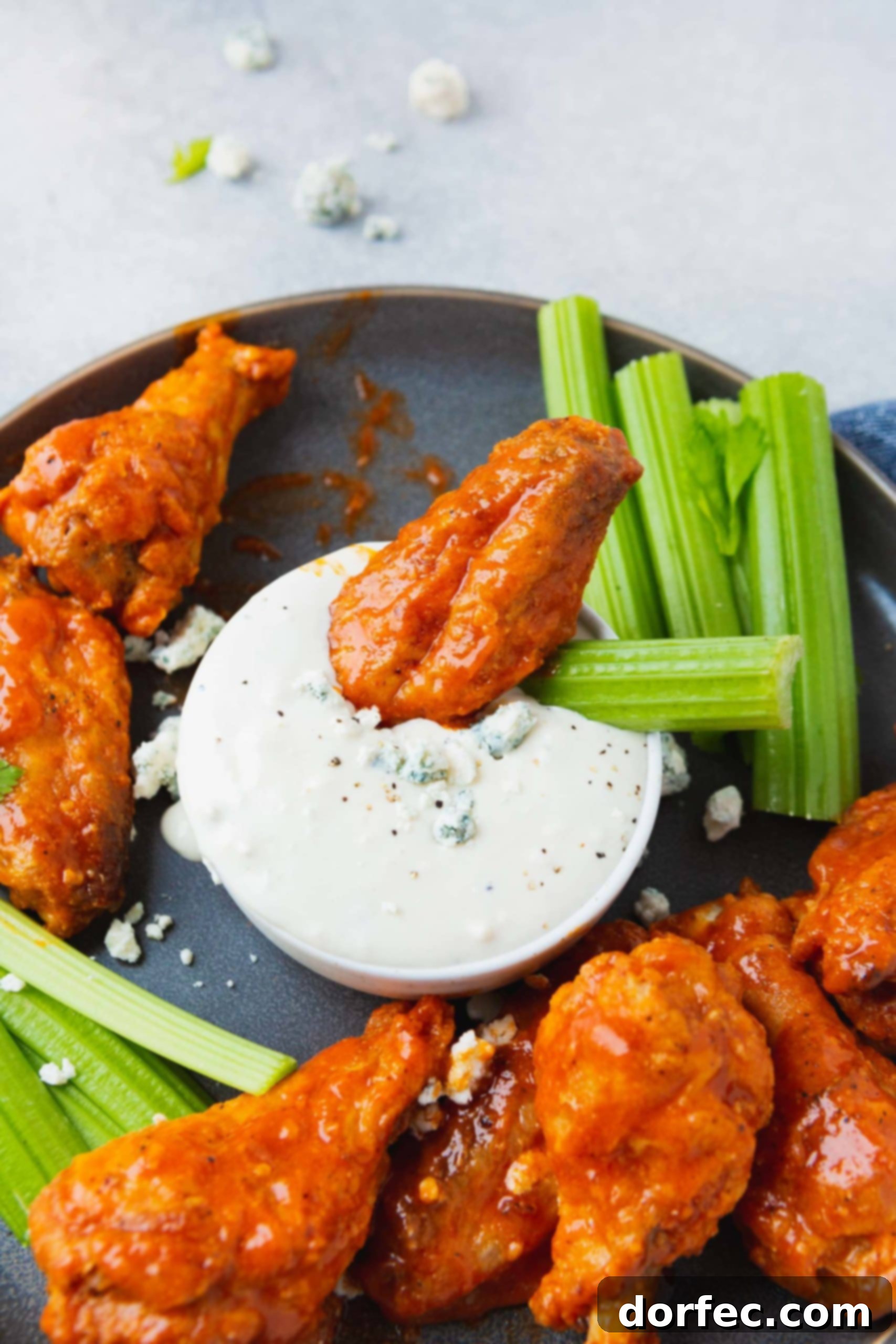 A charcoal platter piled high with crispy buffalo wings and fresh celery sticks, with a bowl of creamy blue cheese dressing at the center. One wing and a celery stick are dipped into the dressing.
