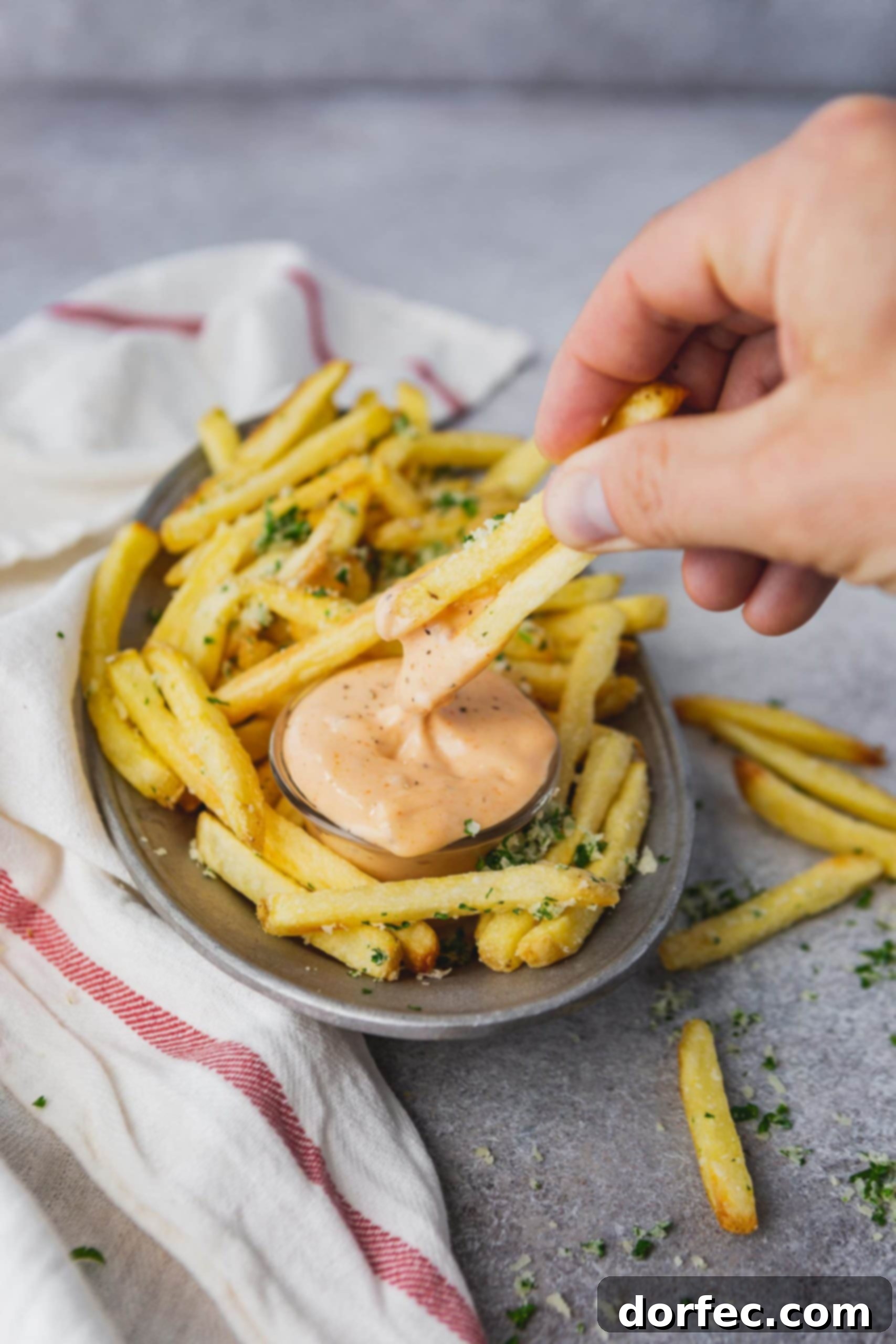 A plate of golden pomme frites with a cup of Cajun dipping sauce next to it, and a hand dipping two fries into the sauce, capturing the anticipation of a delicious bite.