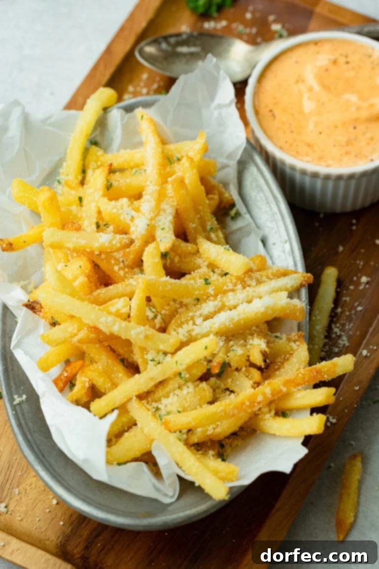 A plate generously piled with hot, salty pomme frites covered in grated Parmesan cheese, nestled beside a small bowl brimming with flavorful Cajun dipping sauce.