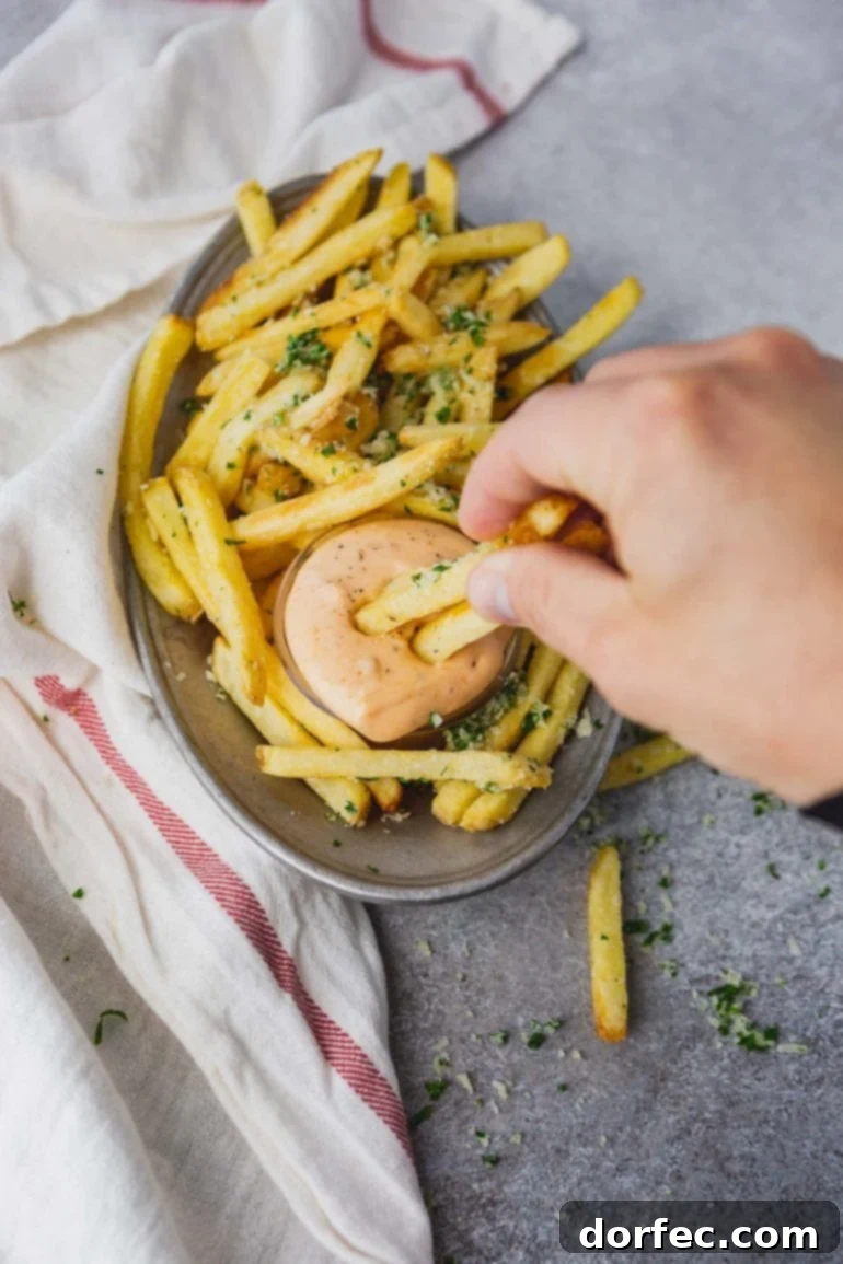 A plate filled with golden pomme frites and a cup of Cajun dipping sauce, with someone actively dipping two fries into the creamy sauce, ready to enjoy.