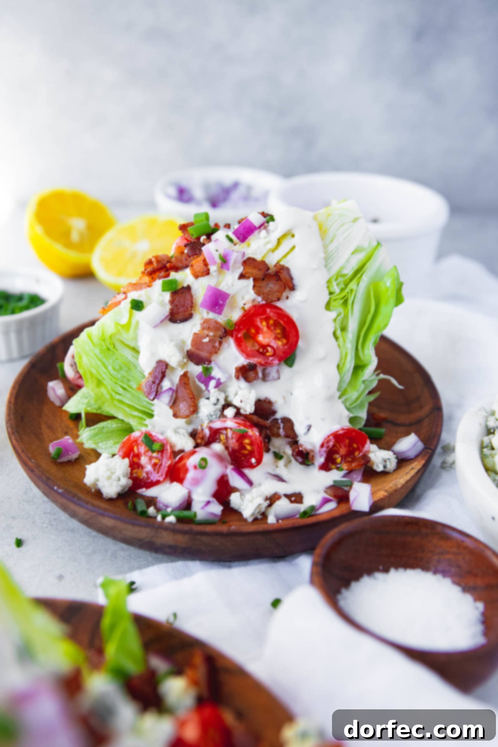 a photo of a wedge salad on a wooden salad plate topped with blue cheese dressing, bacon crumbles, diced red onion, halved cherry tomatoes and blue cheese crumbles.