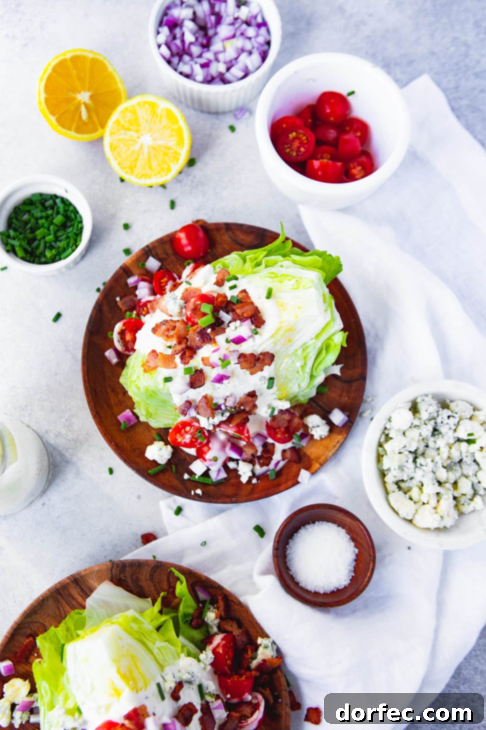 a photo of a wedge salad taken from above the salad. the salad is on a wooden salad plate and topped with blue cheese dressing, bacon crumbles, cherry tomatoes, diced red onion. the plate is surrounded by small containers of the different toppings.