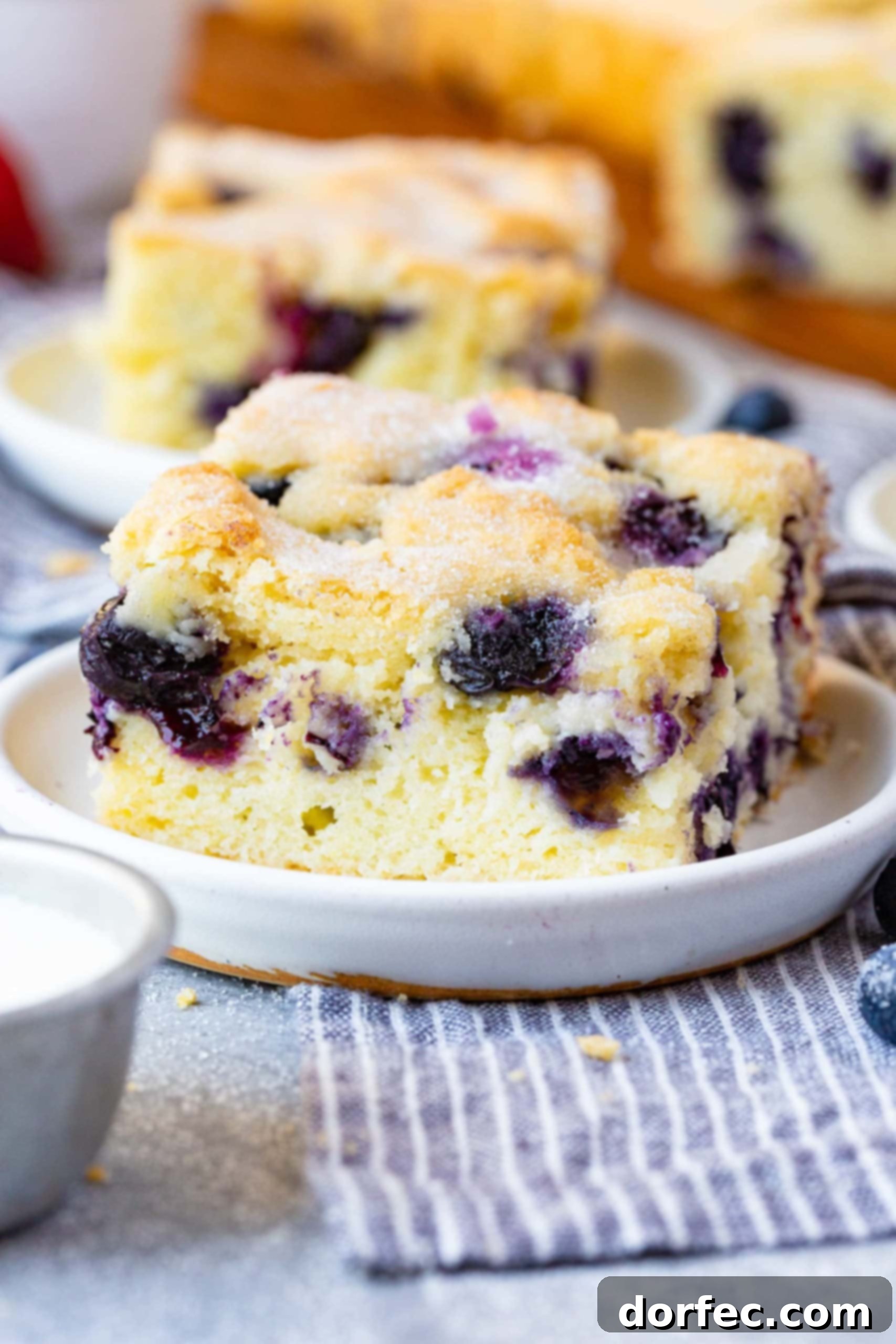 A vibrant slice of buttermilk blueberry breakfast cake, rich with purple fresh blueberries, rests on a pristine white plate, invitingly set against a backdrop of scattered whole blueberries.