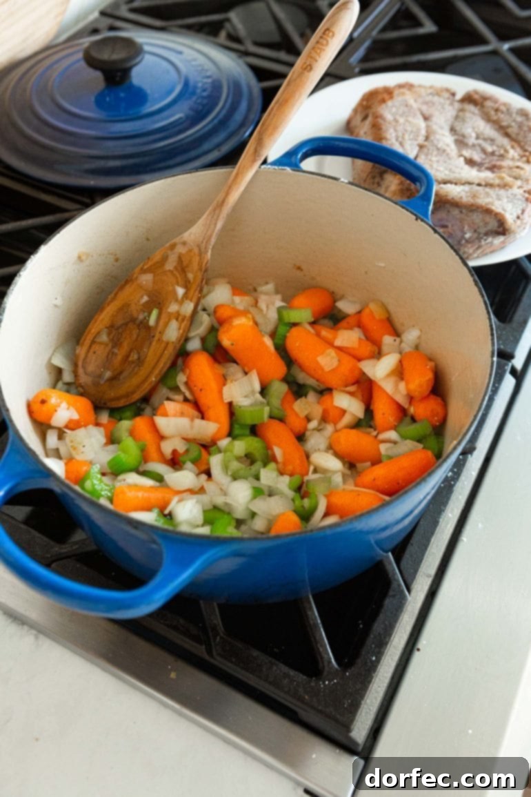 A photo of a large dutch oven will of whole baby carrots, diced onions and celery with a wooden spoon in the pot.