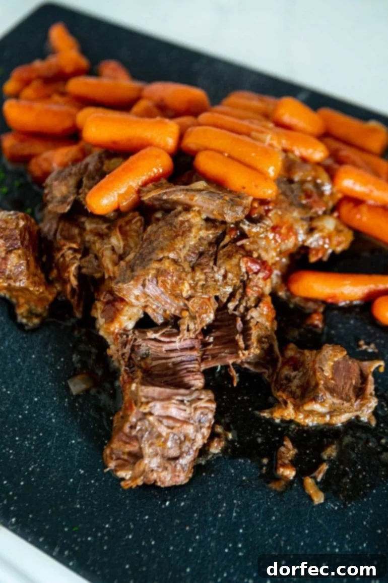 A photo of tender beef roast being shredded on a serving platter with cooked whole baby carrots in the background.