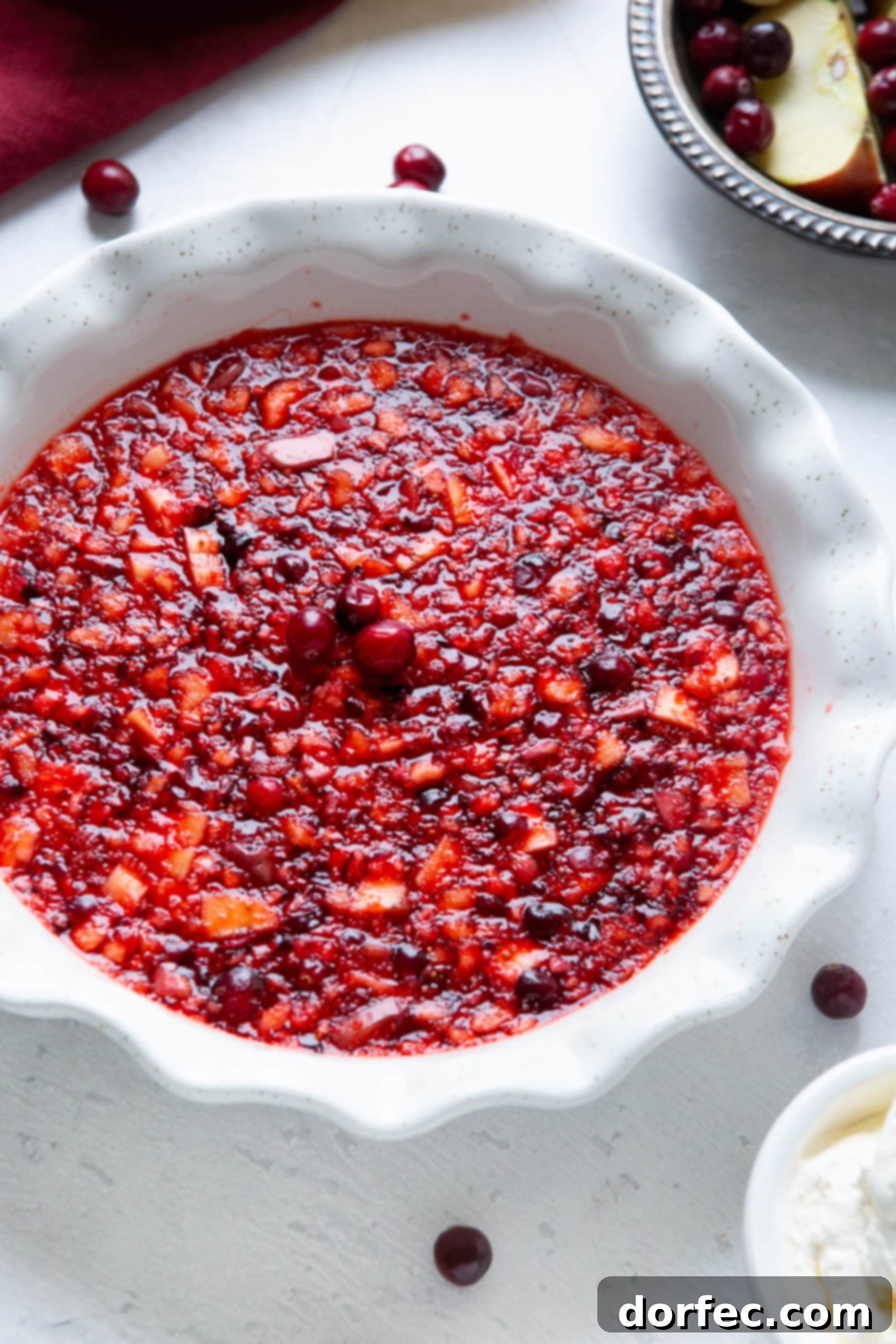 A close-up view of a white ceramic pie dish filled with a chunky, red fruit-filled Jell-O, elegantly garnished with three fresh cranberries.
