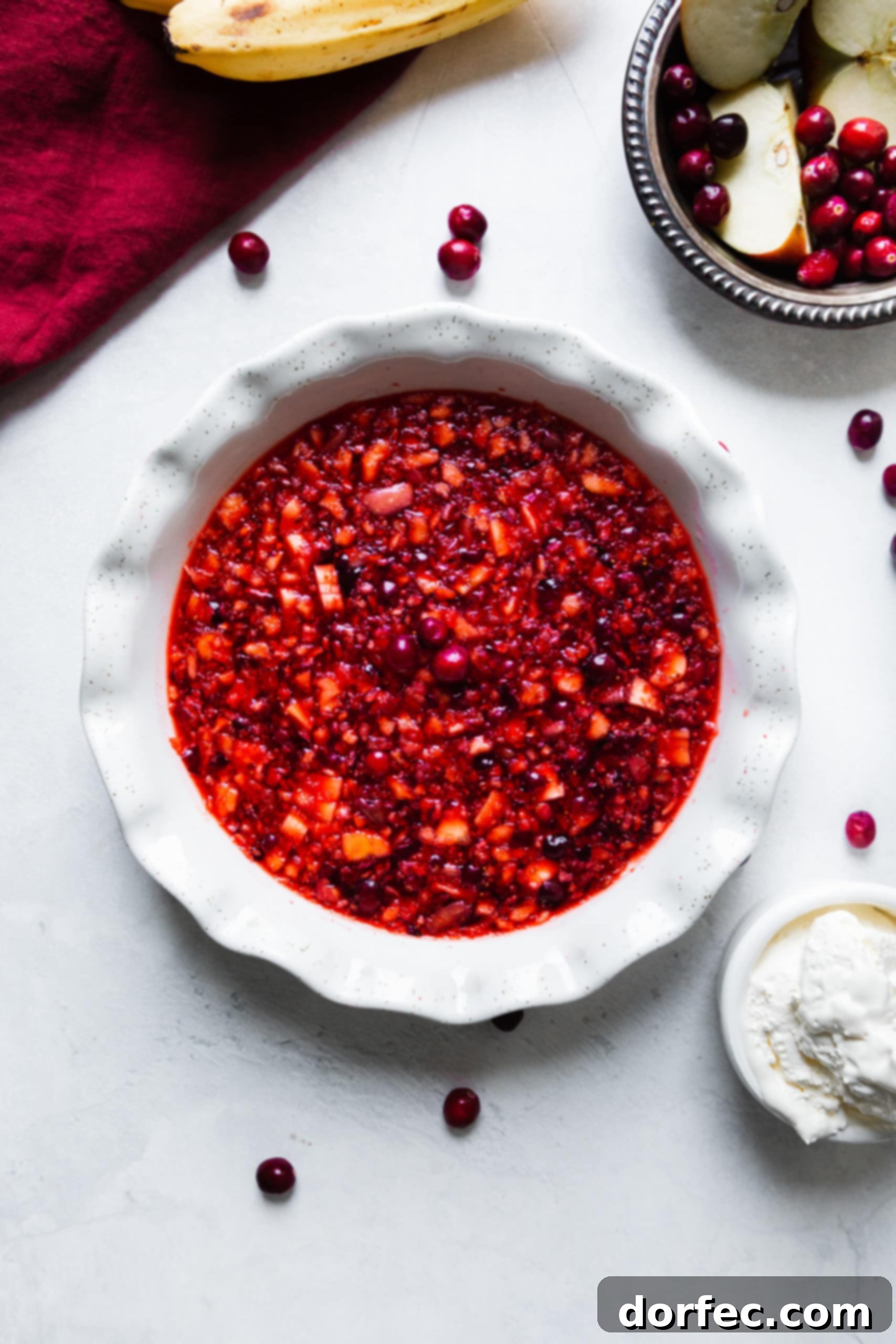 An overhead shot of a white ceramic pie dish filled with chunky fruit Jell-O, accompanied by a small bowl of fresh whipped cream.