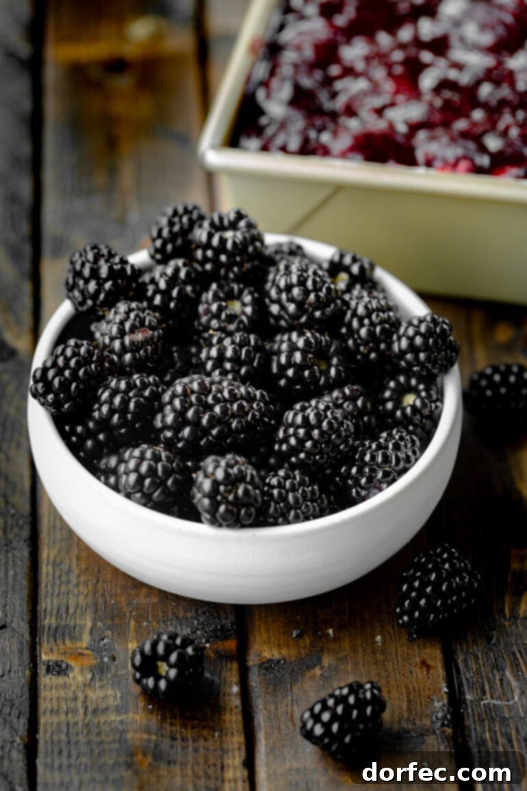 A rustic wooden table displays a bowl overflowing with plump, dark marionberries, with a slice of berry cheesecake bar artfully blurred in the background.