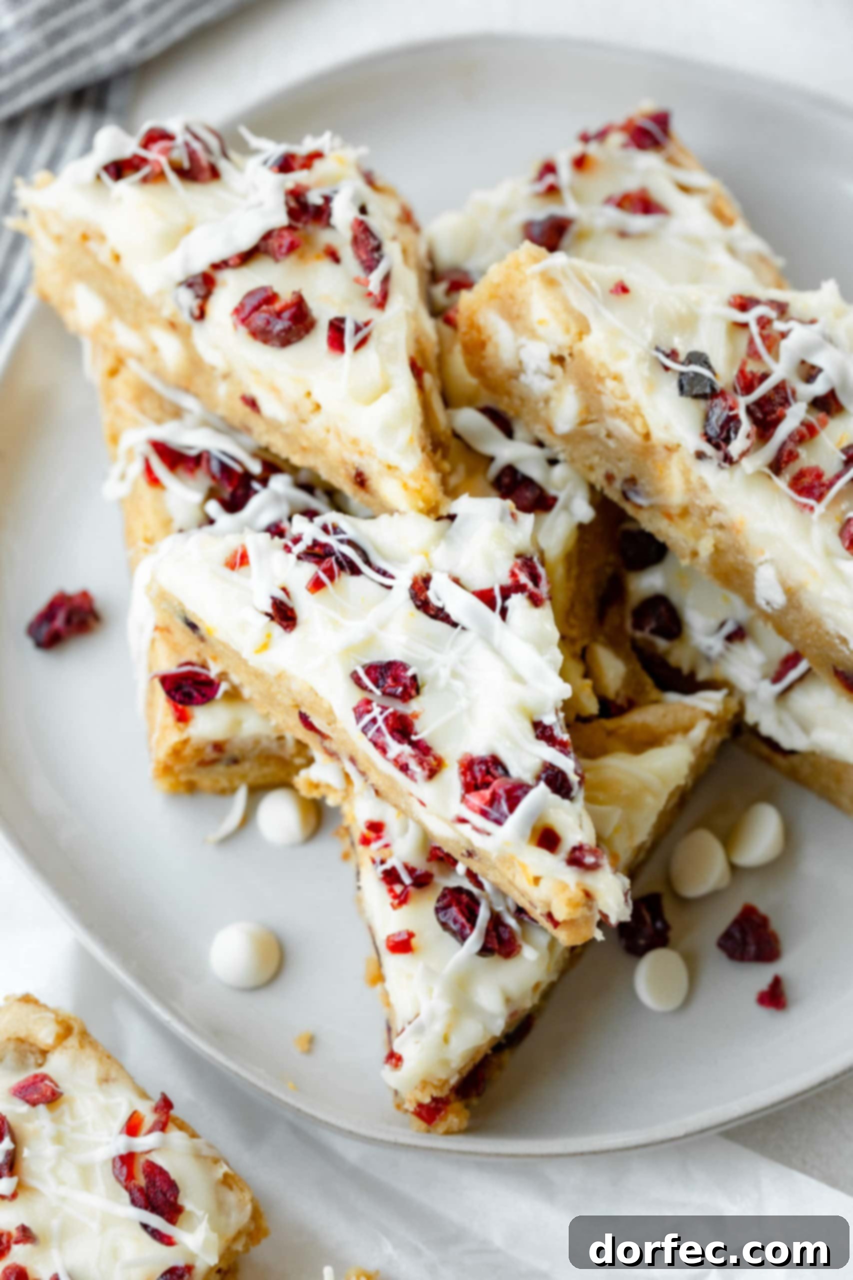 A photo of several orange cranberry bliss bars, cut into triangles and stacked, beautifully drizzled with white chocolate and garnished with dried cranberries.