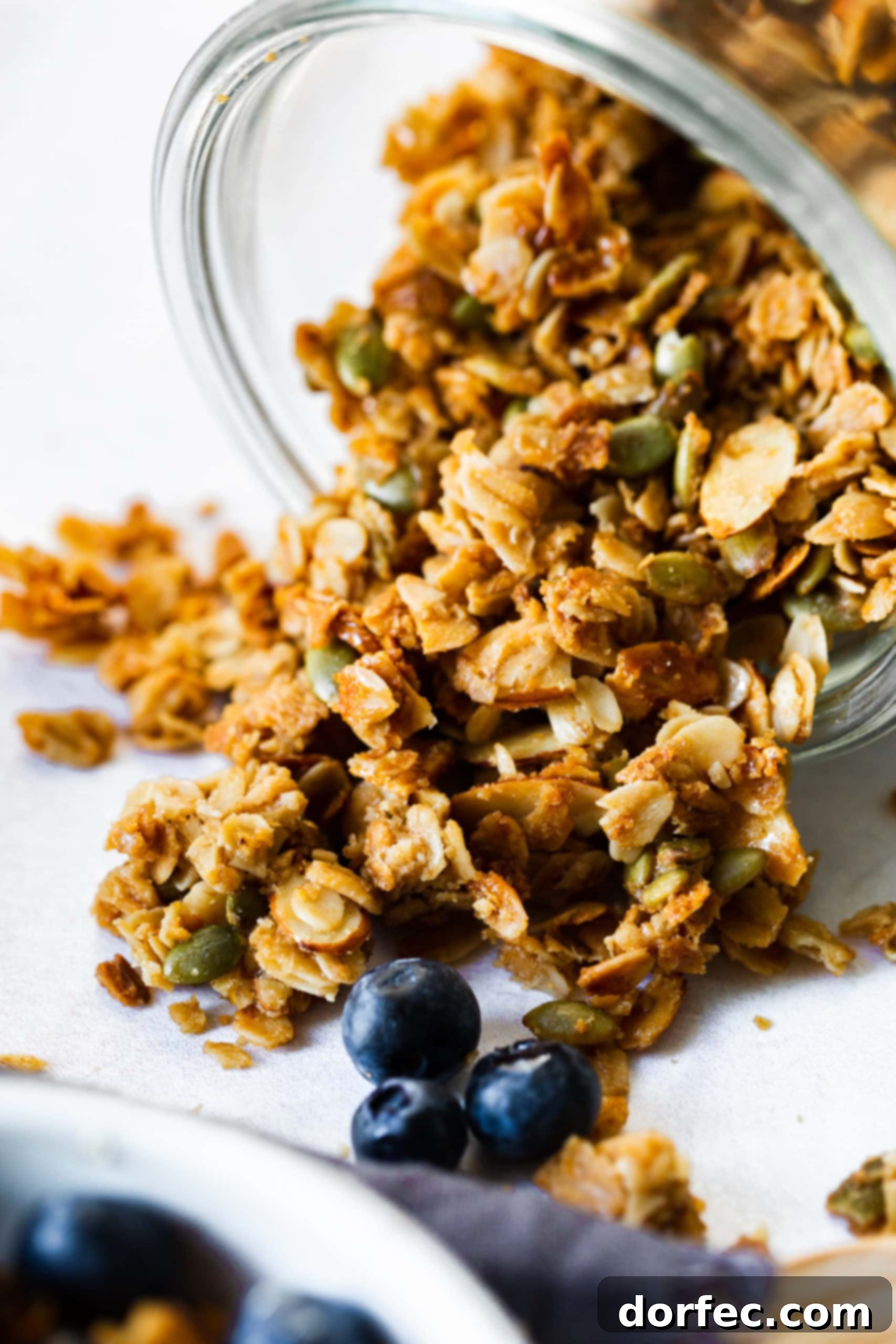 A photo of homemade granola being poured out of a container with fresh blueberries in the foreground.