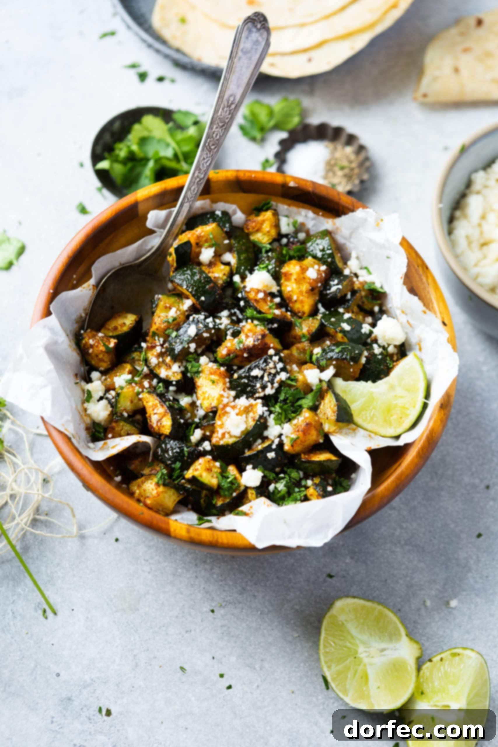 A large wooden serving bowl overflowing with beautifully roasted zucchini, garnished with crumbled queso fresco and a fresh lime wedge on the side.
