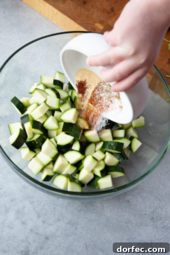 All the vibrant Mexican seasonings being poured into a large glass bowl with bite-sized zucchini chunks, ensuring every piece is coated for maximum flavor.