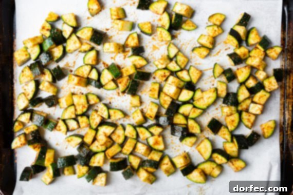 Seasoned raw zucchini chunks spread evenly on white parchment paper on a baking sheet, ready for the oven to transform them.