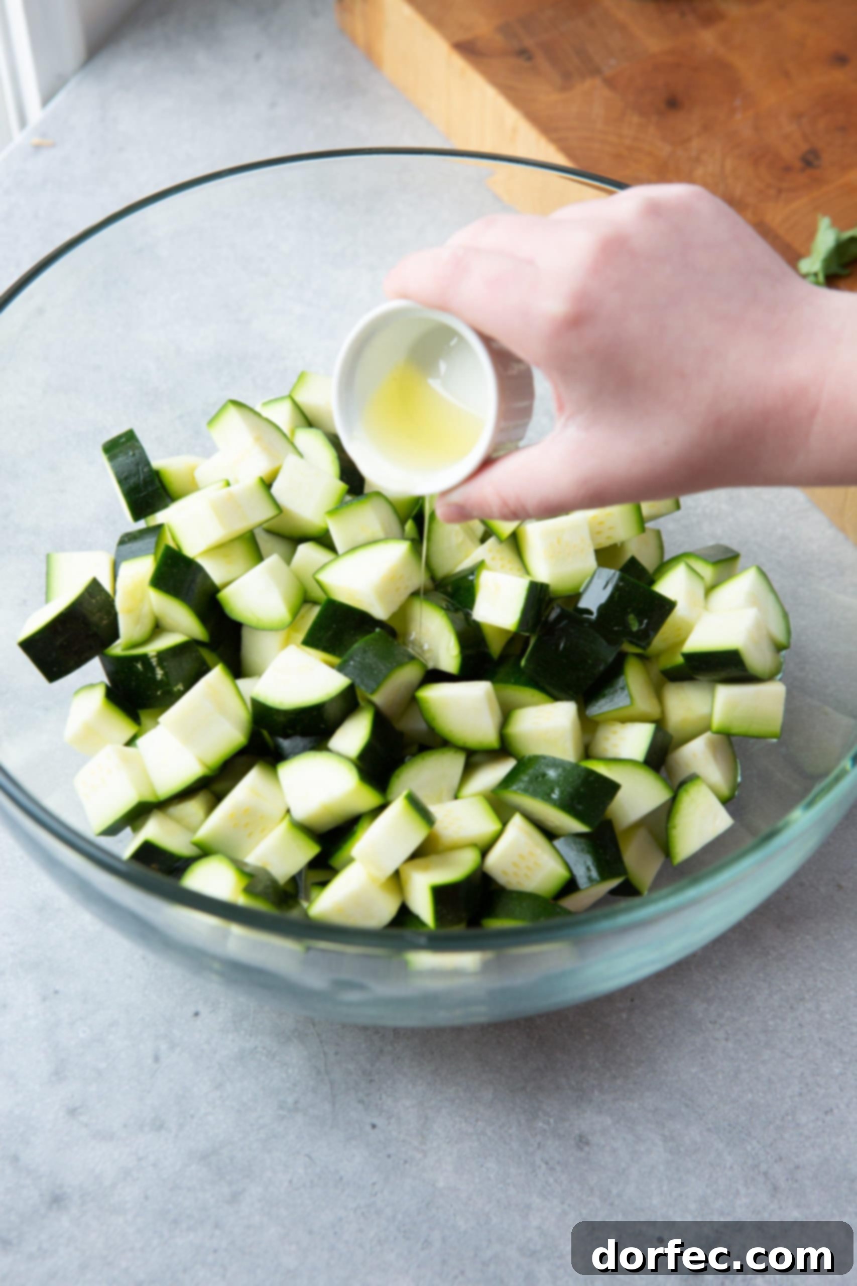 Someone pouring olive oil onto bite-sized zucchini chunks in a large glass mixing bowl, a crucial step before seasoning.