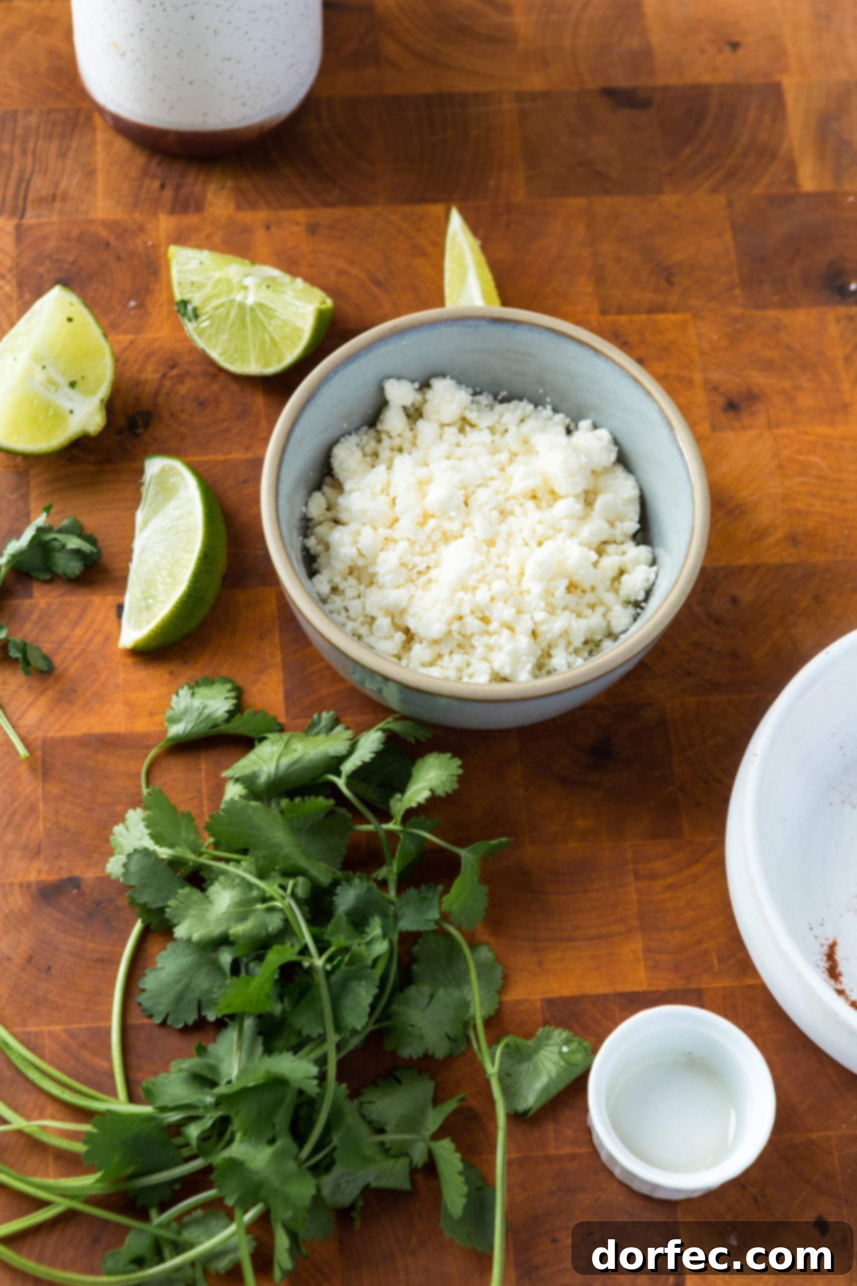 A selection of fresh toppings for Mexican roasted zucchini: a bunch of fresh cilantro, four lime wedges, and a small bowl of crumbled queso fresco, all arranged on a wooden cutting board.