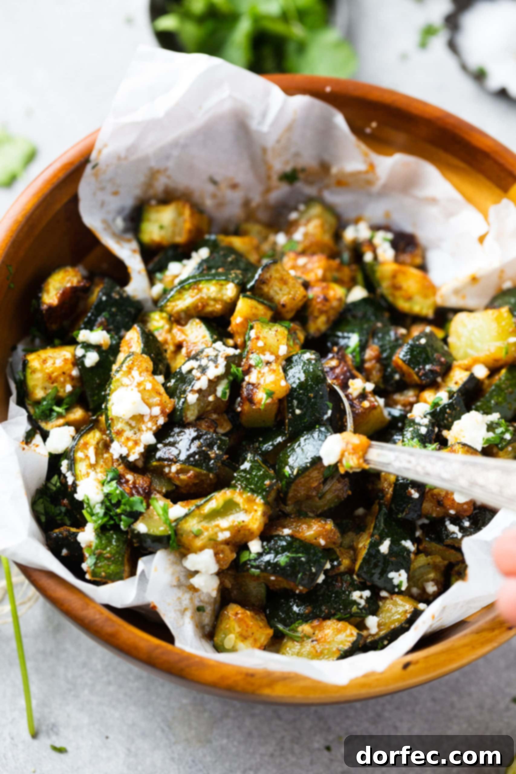 A large silver serving spoon scooping out a generous portion of Mexican roasted zucchini, beautifully topped with crumbled queso fresco, from a wooden bowl.