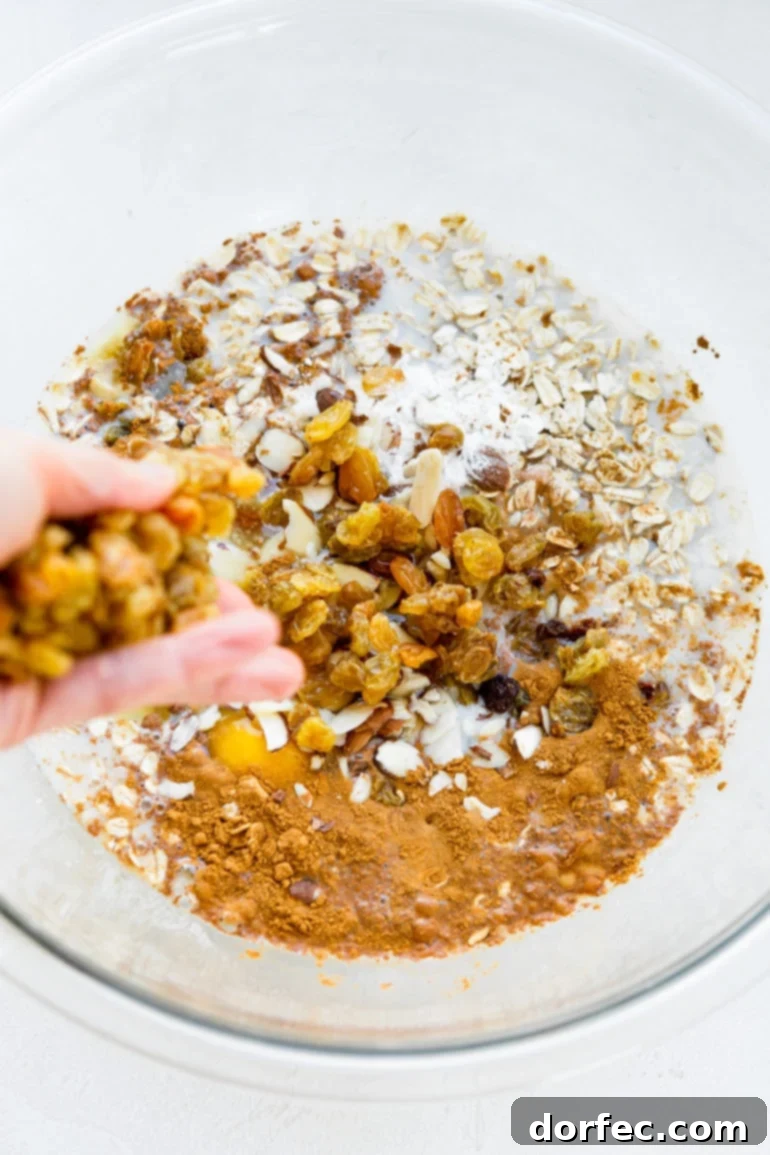 Baked Apple Fritter Oatmeal Delight 4 A photo of a hand dropping golden raisins into a large glass bowl, which is already filled with uncooked oatmeal, dried apple chunks, and spices.