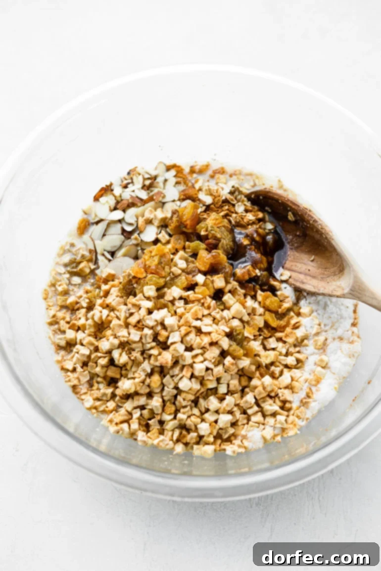 Baked Apple Fritter Oatmeal Delight 5 A photo of a wooden spoon stirring a large glass bowl filled with uncooked oatmeal, dried apple chunks, and spices, combining all the ingredients.