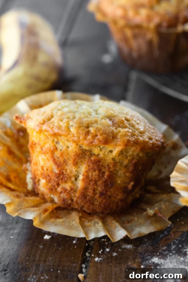 Close-up photo of a baked banana muffin with the paper wrapper partially peeled off, revealing its soft, golden interior.