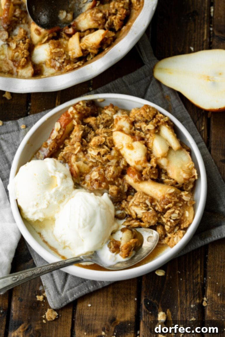 A close-up shot of a bowl of pear crisp, showing tender, juicy pears beneath a golden, crunchy oat topping, with two scoops of vanilla ice cream on the side.