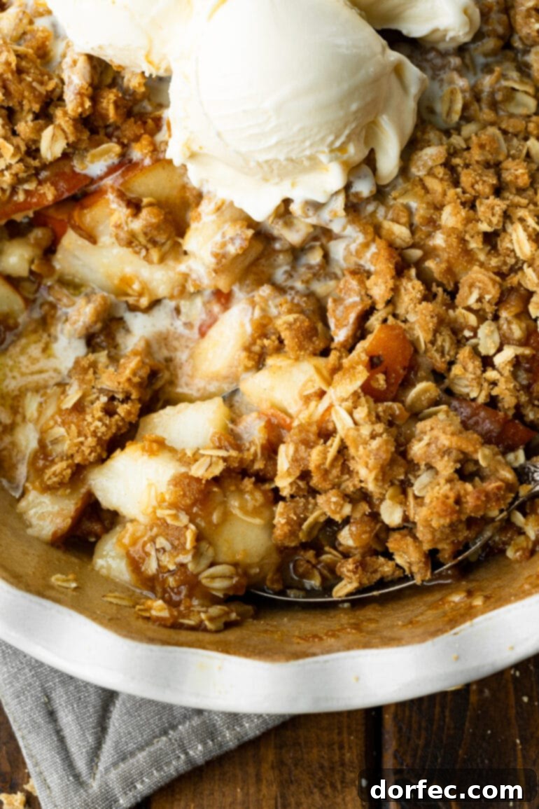 A circular baking dish showcasing a beautifully baked pear crisp with a golden, bubbly oat topping, served with scoops of melting vanilla ice cream.