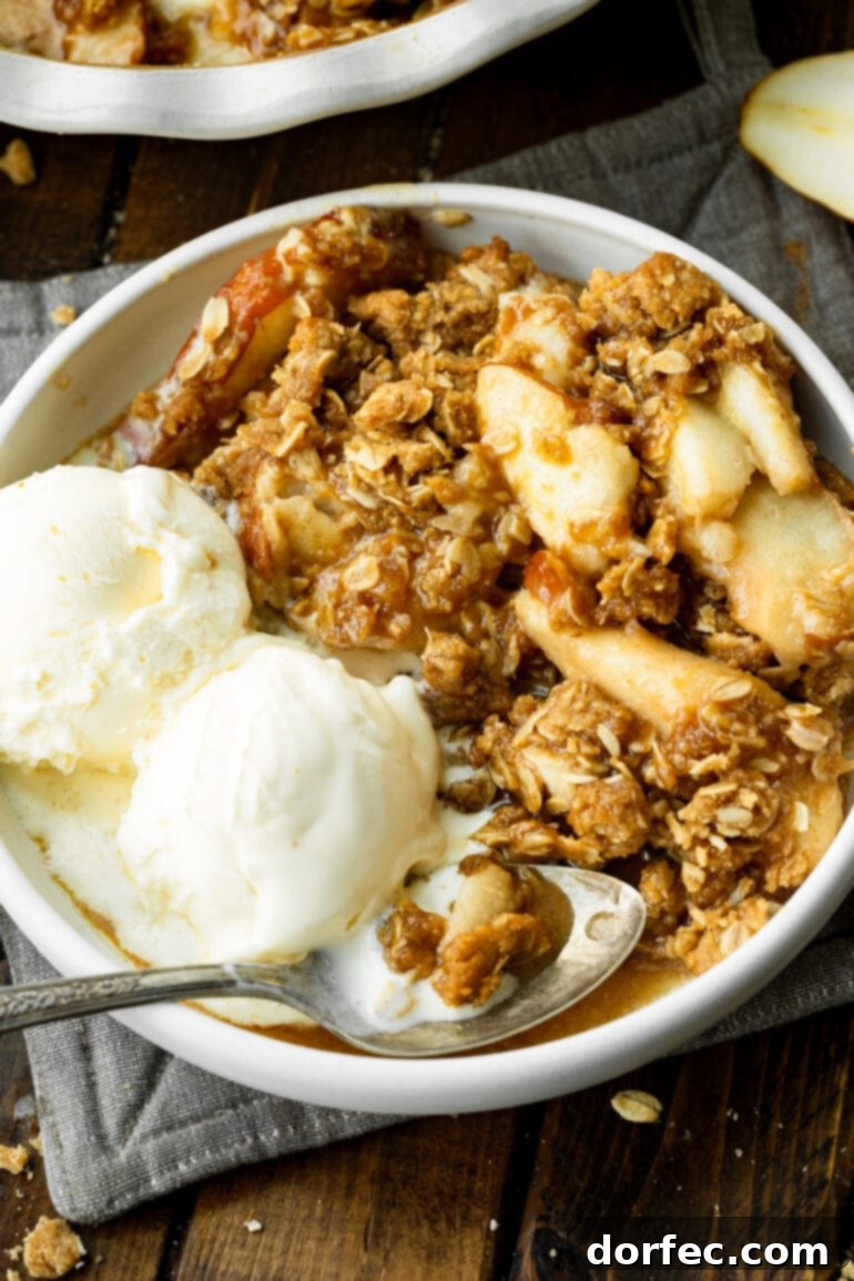 A close-up of a bowl of pear crisp, showing the tender, juicy pear filling beneath a golden, crunchy oat topping, with two scoops of vanilla ice cream on the side.