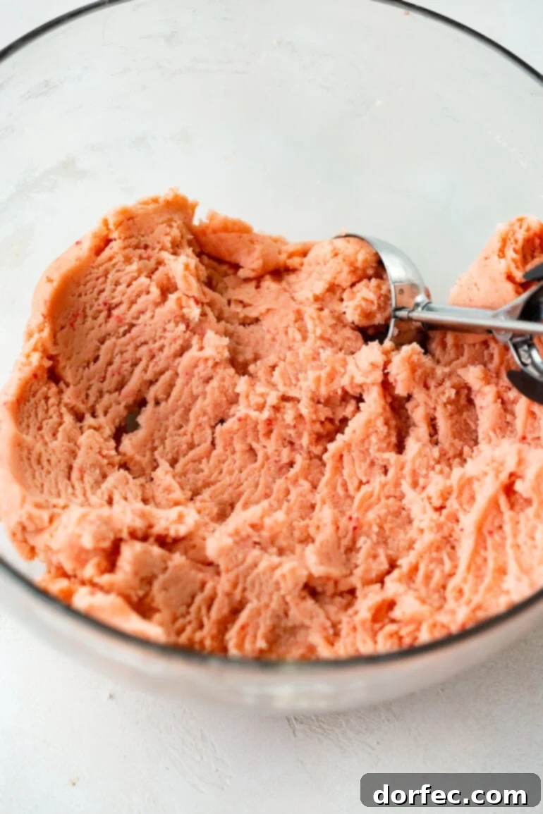 A candid photo showing a large, clear glass bowl brimming with subtly pink cookie dough. A cookie scoop is actively scooping out portions, ready to be shaped and baked. The natural, delicate pink color of the dough is beautifully visible.