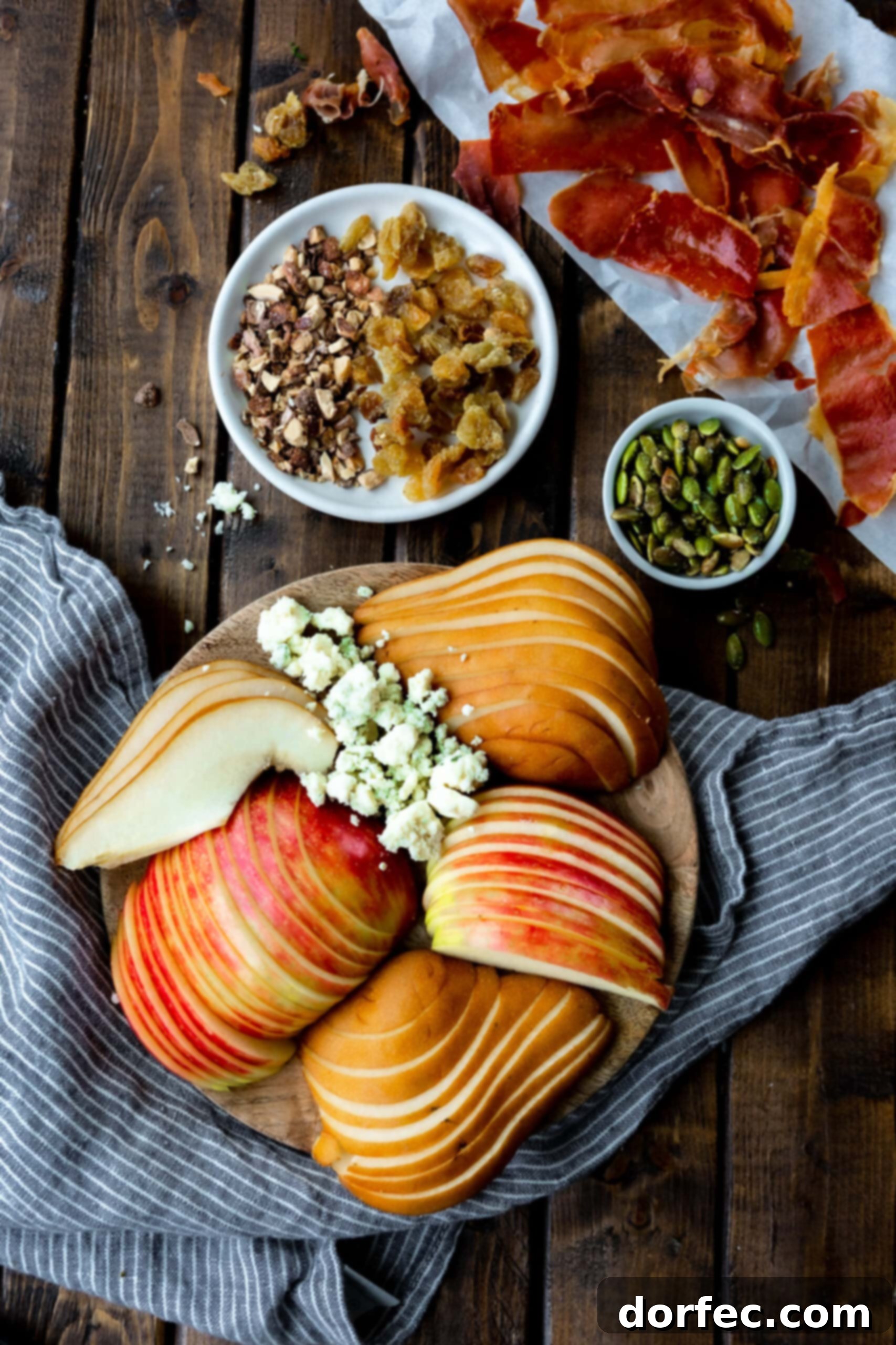 Fresh Ingredients for Roasted Pear & Apple Salad A beautifully composed flat lay of thinly sliced apples and pears, ready for roasting, alongside crumbled gorgonzola cheese, a small bowl of pepitas, and golden raisins with chopped cinnamon pecans, highlighting the fresh ingredients for the salad.
