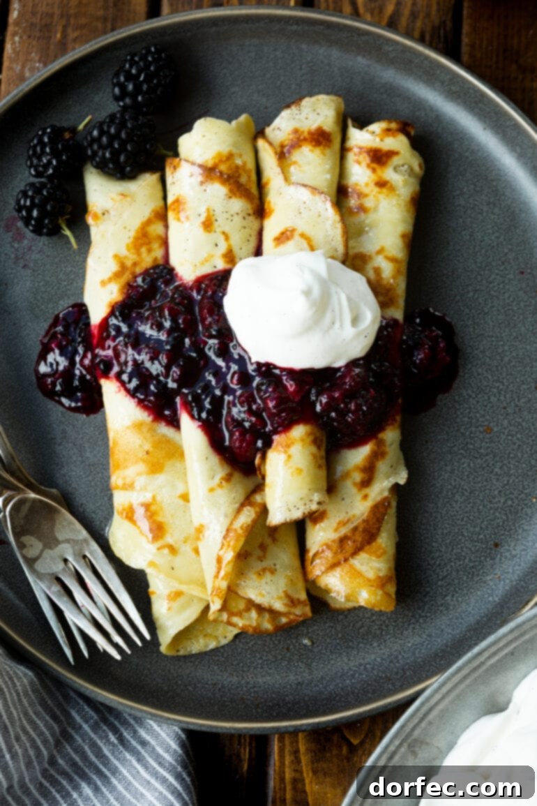 a photo of several rolled up finnish pancakes sitting on a dark gray plate and topped with blackberry syrup and a dollop of whipped cream.
