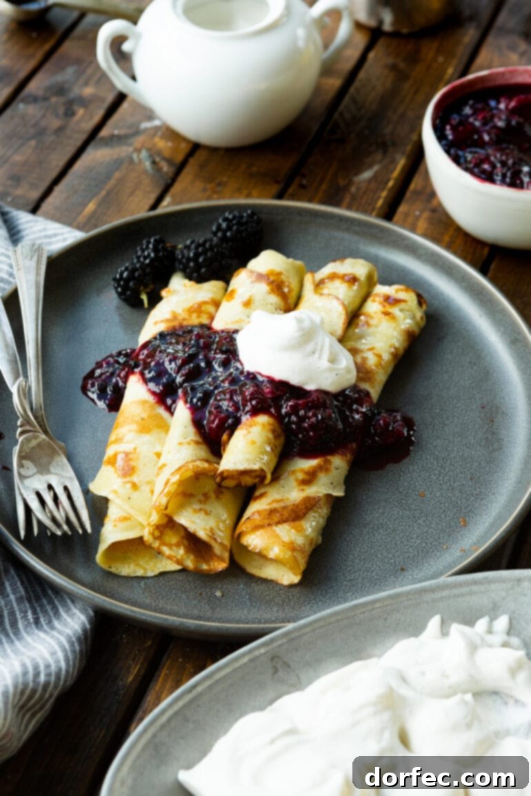 a photo of five rolled up golden finnish pancakes topped with a blackberry sauce and a dollop of whipped cream.